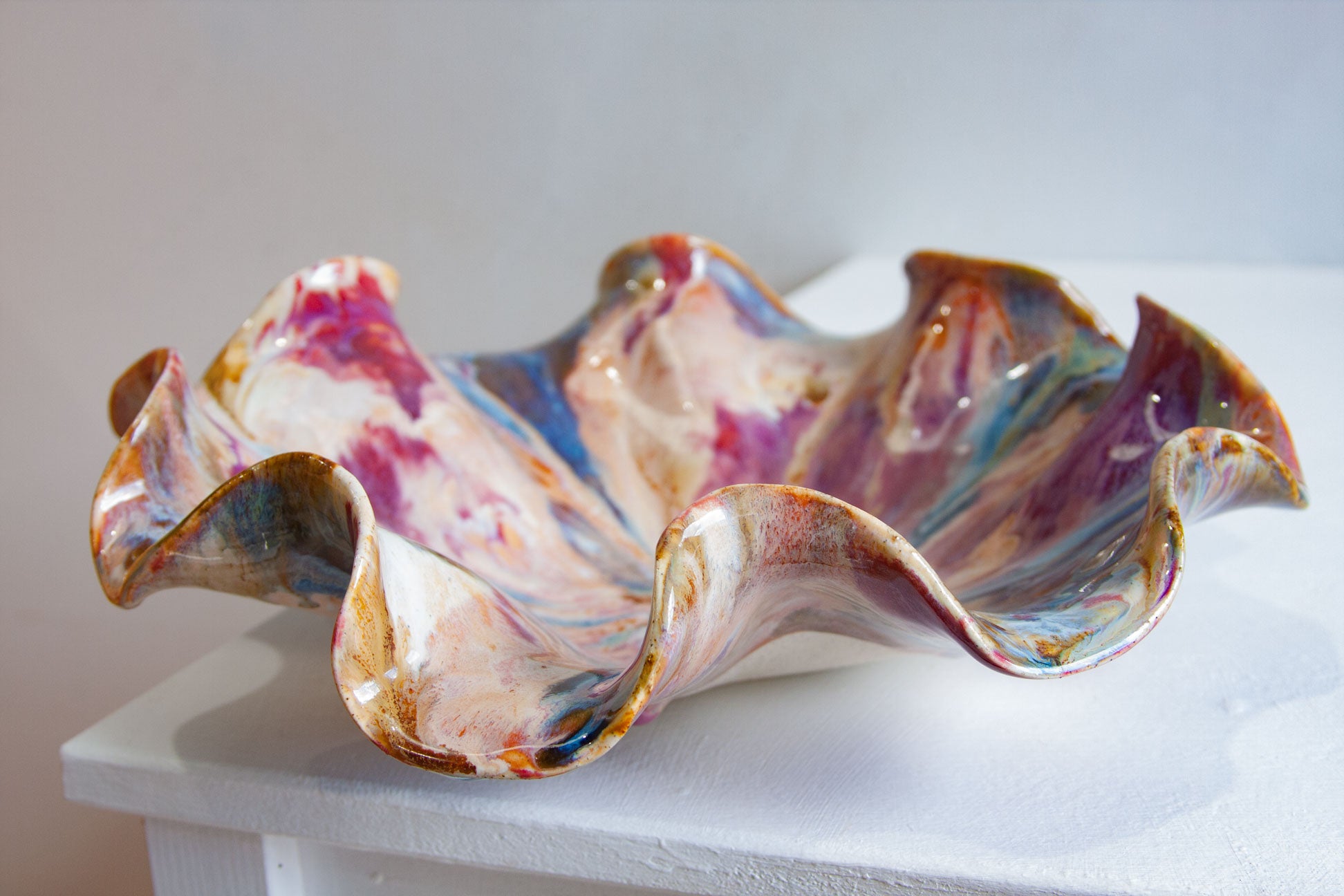 A handmade ceramic bowl with a wavy, ruffled edge, decorated with a marbled glaze of pink, purple, blue, gold, and white, displayed on a white pedestal against a neutral gray background.