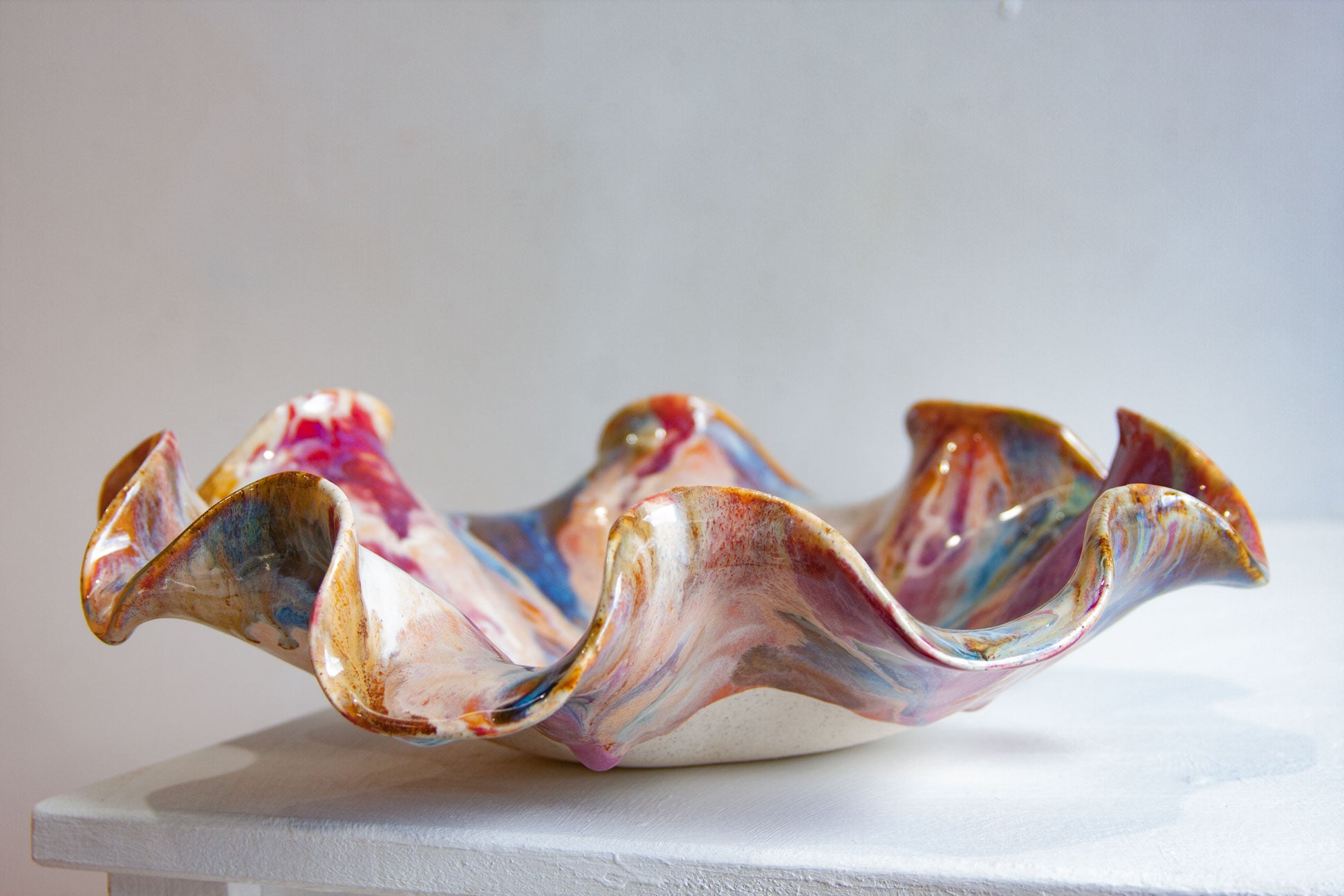 A handmade ceramic bowl with a wavy, ruffled rim, decorated with a marbled glaze of pink, blue, brown, and white, displayed on a white surface against a light gray background.