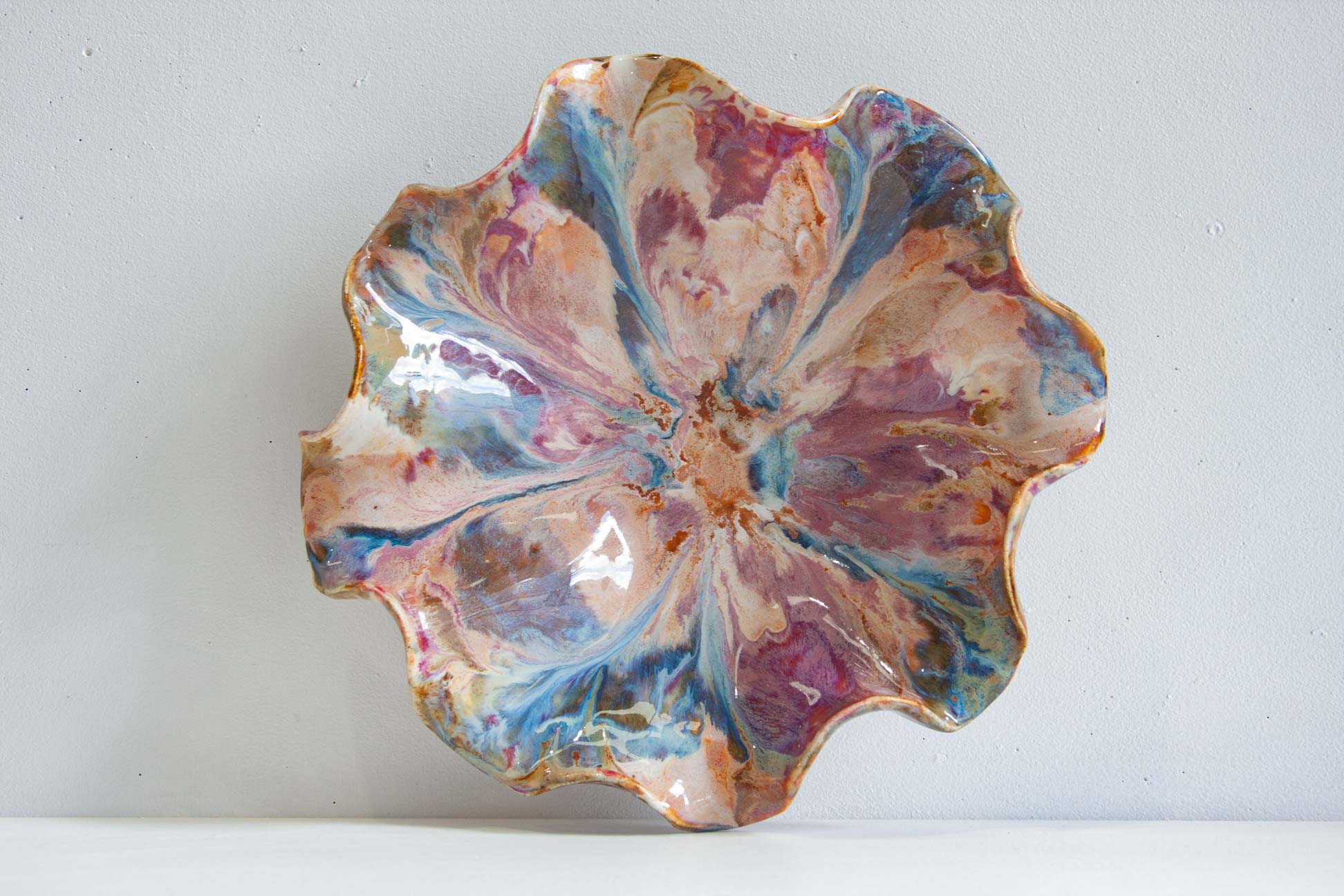 A scalloped ceramic plate with a marbled floral pattern in pink, blue, purple, and gold, photographed against a neutral light gray background.