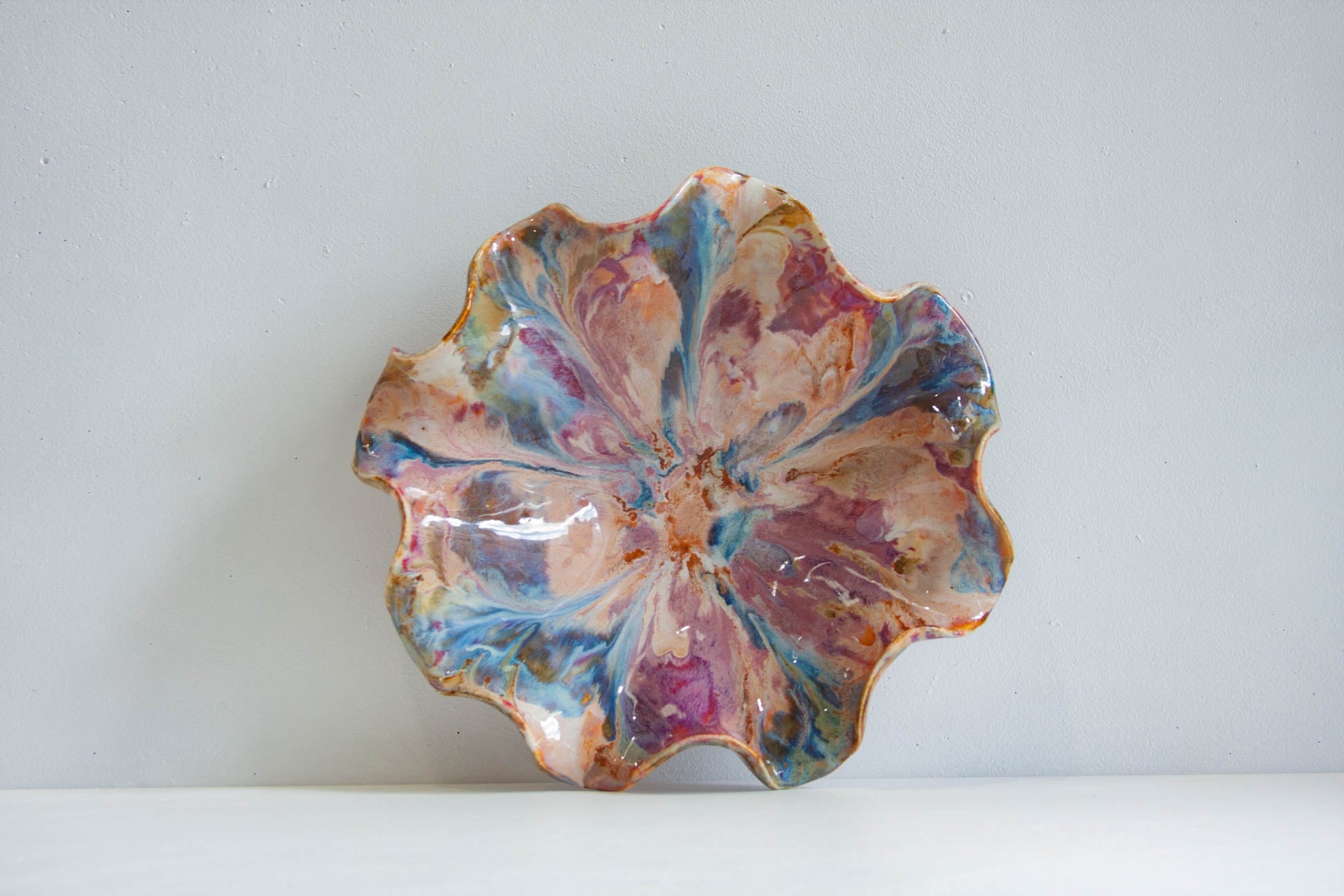 A handmade ceramic flower-shaped bowl with a marbled, swirled glaze in shades of pink, blue, purple, and gold, set against a plain light gray background.