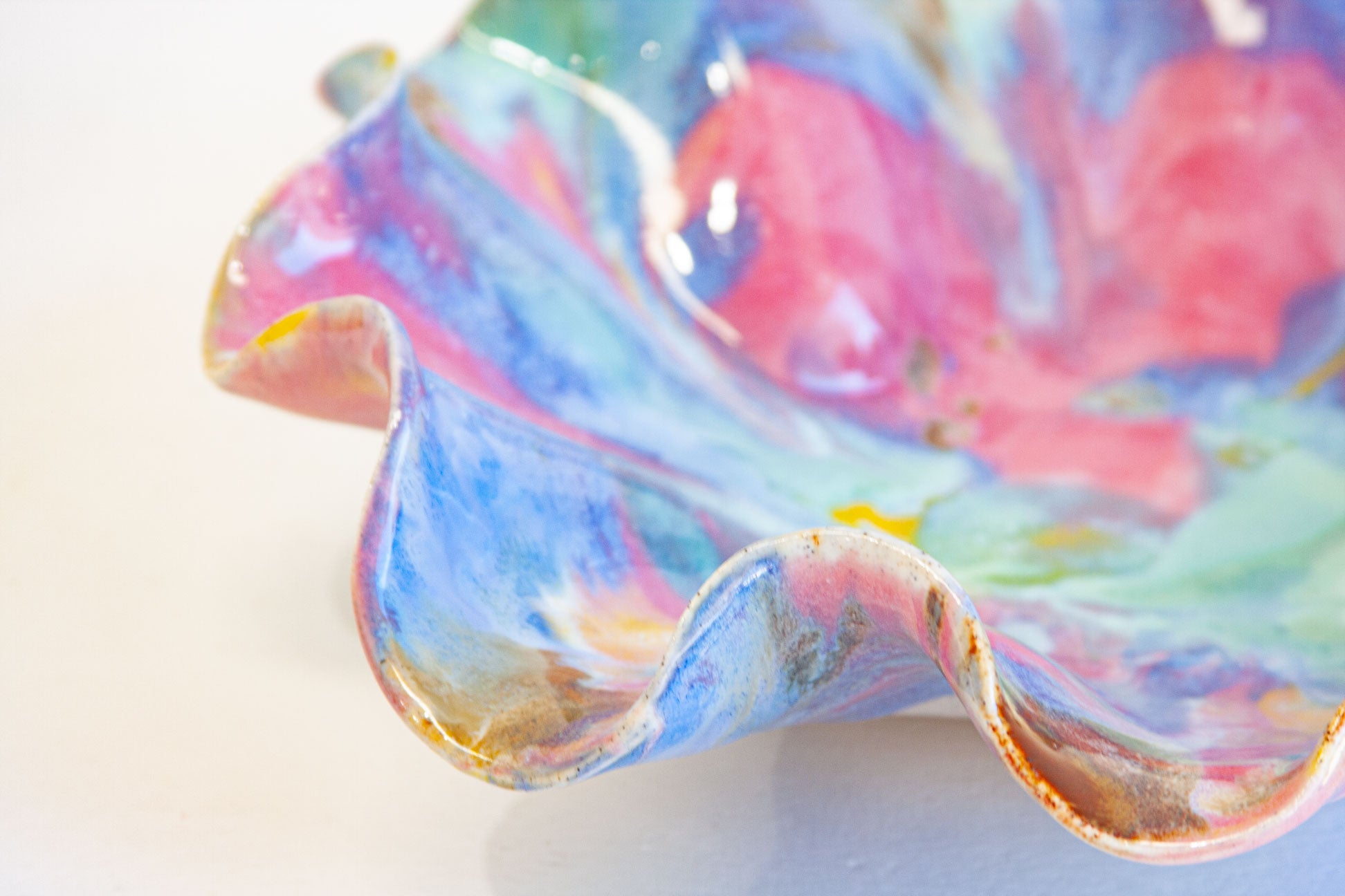 A close-up of a handmade ceramic bowl with a wavy rim, decorated in a marbled pattern of pink, blue, green, and gold glaze on a white background.