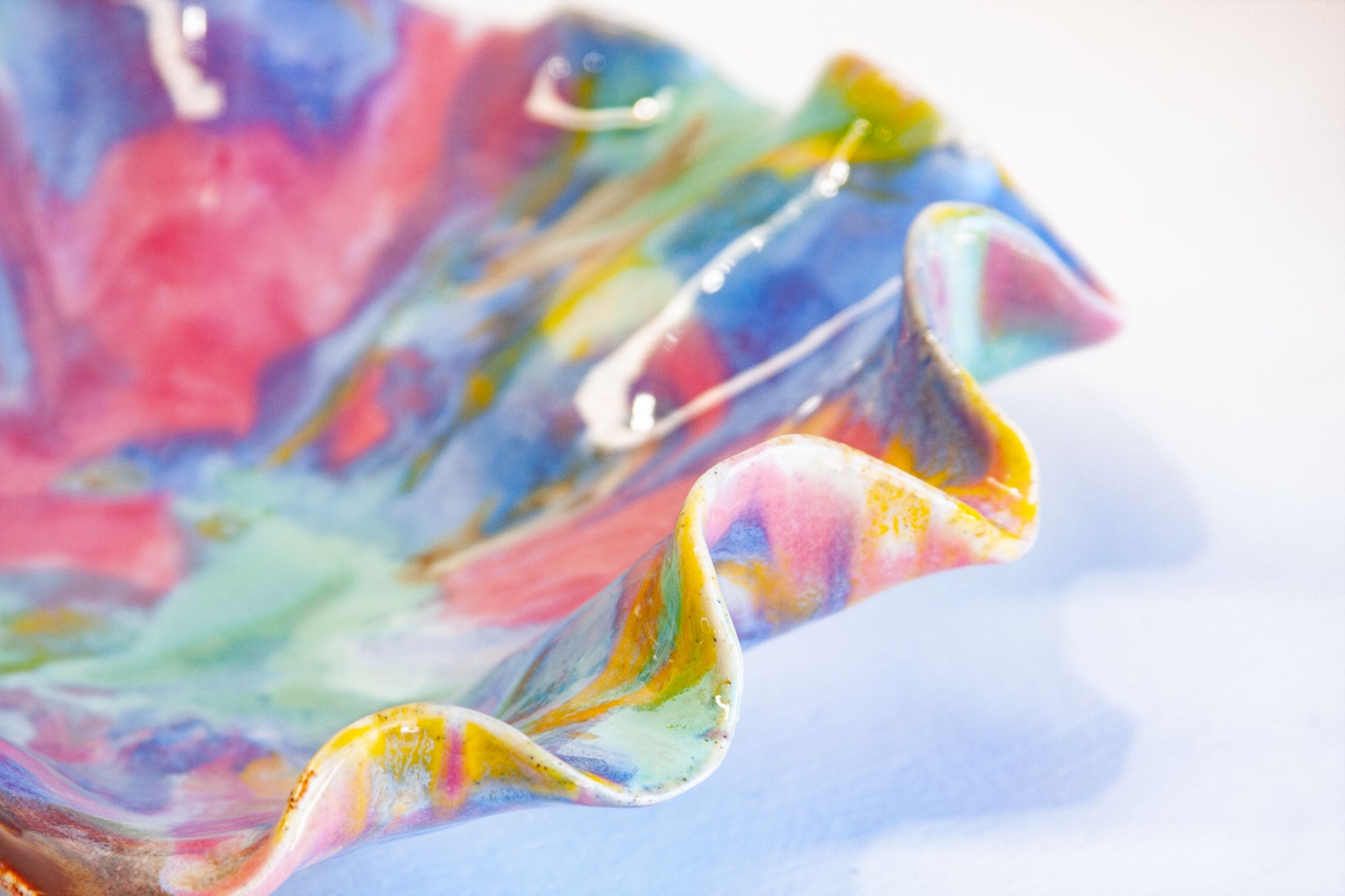 A close-up of a colorful, marbled ceramic dish with a wavy, ruffled rim. The glossy glaze features swirls of pink, blue, green, yellow, and purple on a white background.