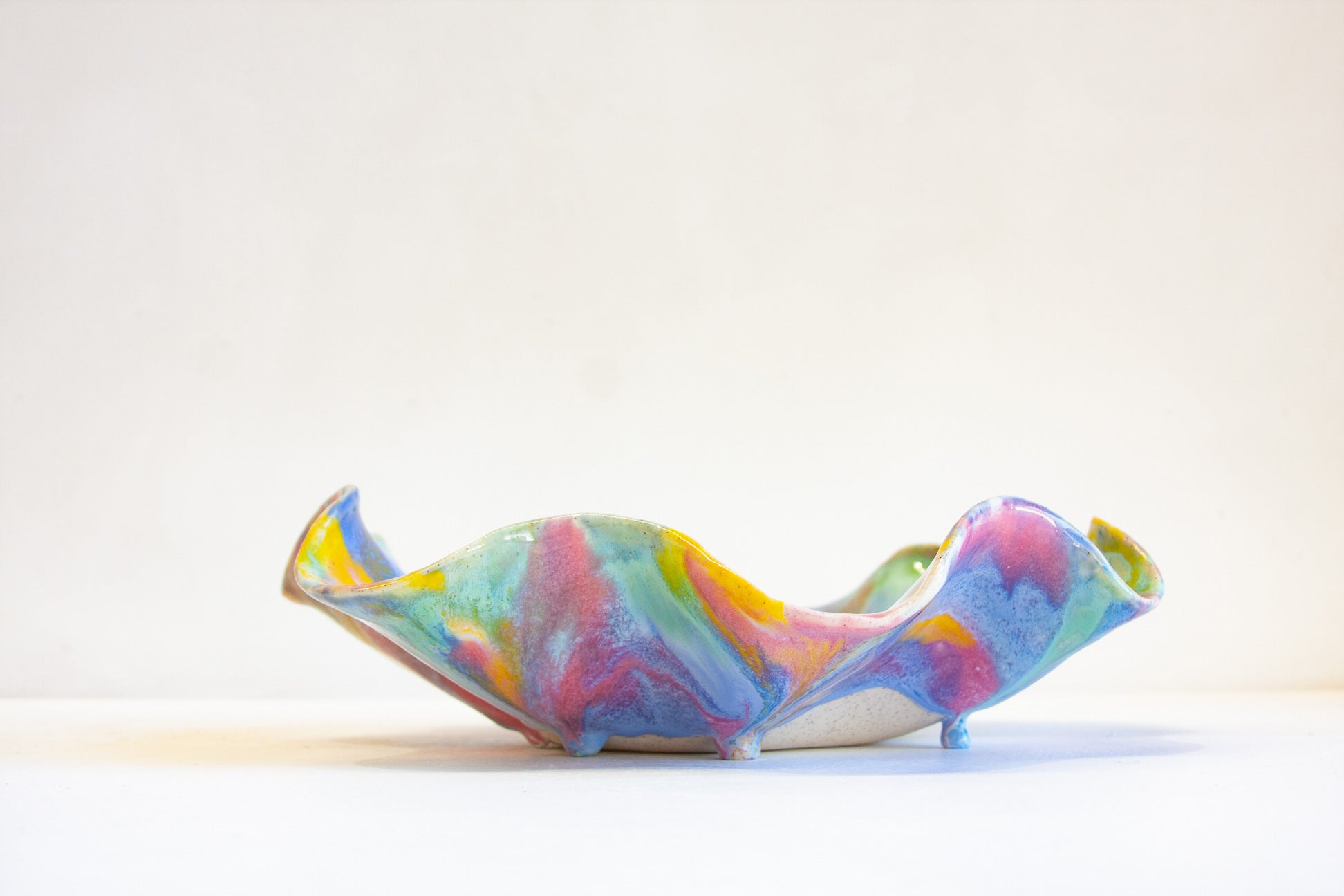 A colorful, sculptural ceramic dish with a marbled swirl pattern in pink, blue, green, yellow, and purple, set against a plain off-white background.