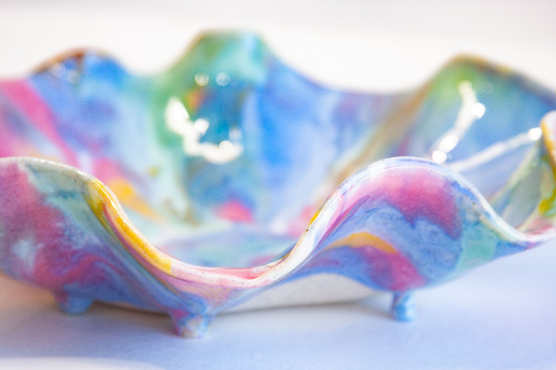 A close-up of a small ceramic dish with a wavy, scalloped rim and three short feet, decorated in a marbled swirl of pastel blue, pink, purple, green, and yellow.