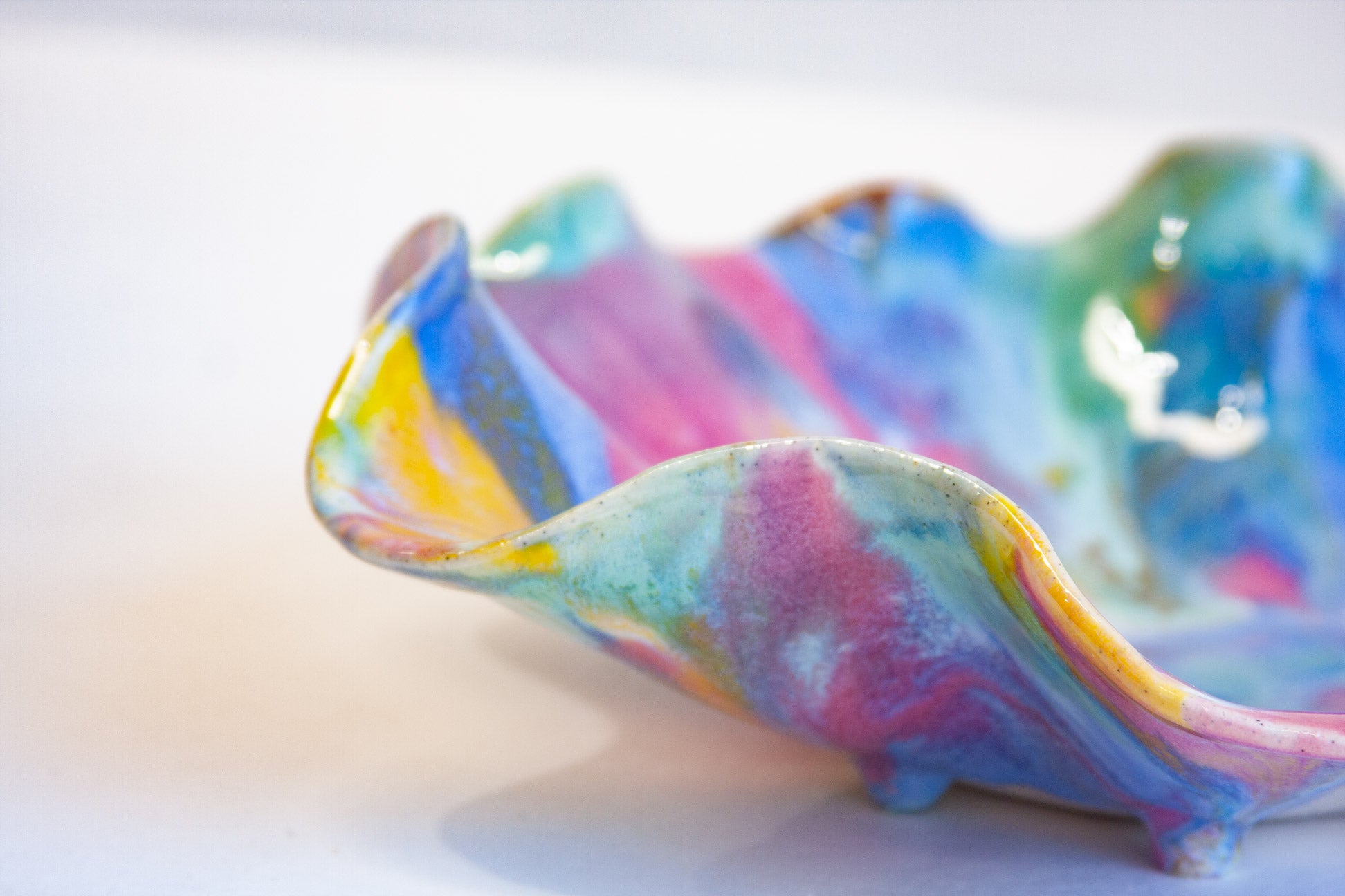 A close-up of a wavy ceramic dish with a marbled, iridescent glaze in pastel blue, pink, yellow, and green on a white background.