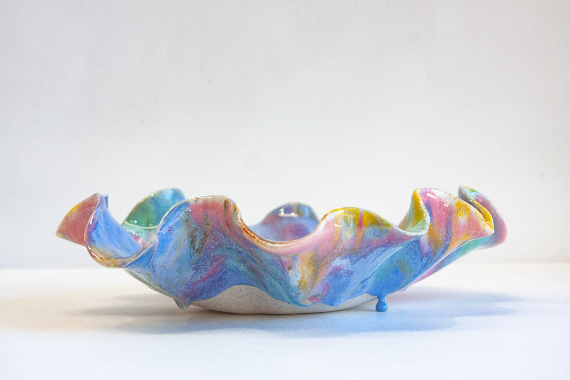 A handmade ceramic bowl with a wavy, ruffled rim, decorated in a marbled pastel pattern of pink, blue, purple, and yellow glaze over a white base, set against a plain white background.