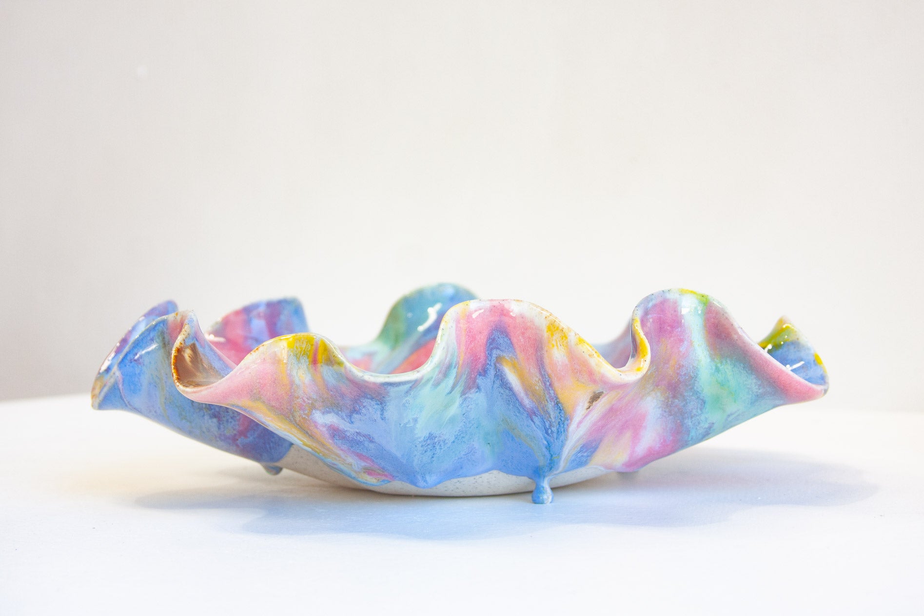 A handmade ceramic dish with a wavy, ruffled rim, decorated in a marbled pattern of pastel blue, pink, purple, and yellow, set against a plain white background.