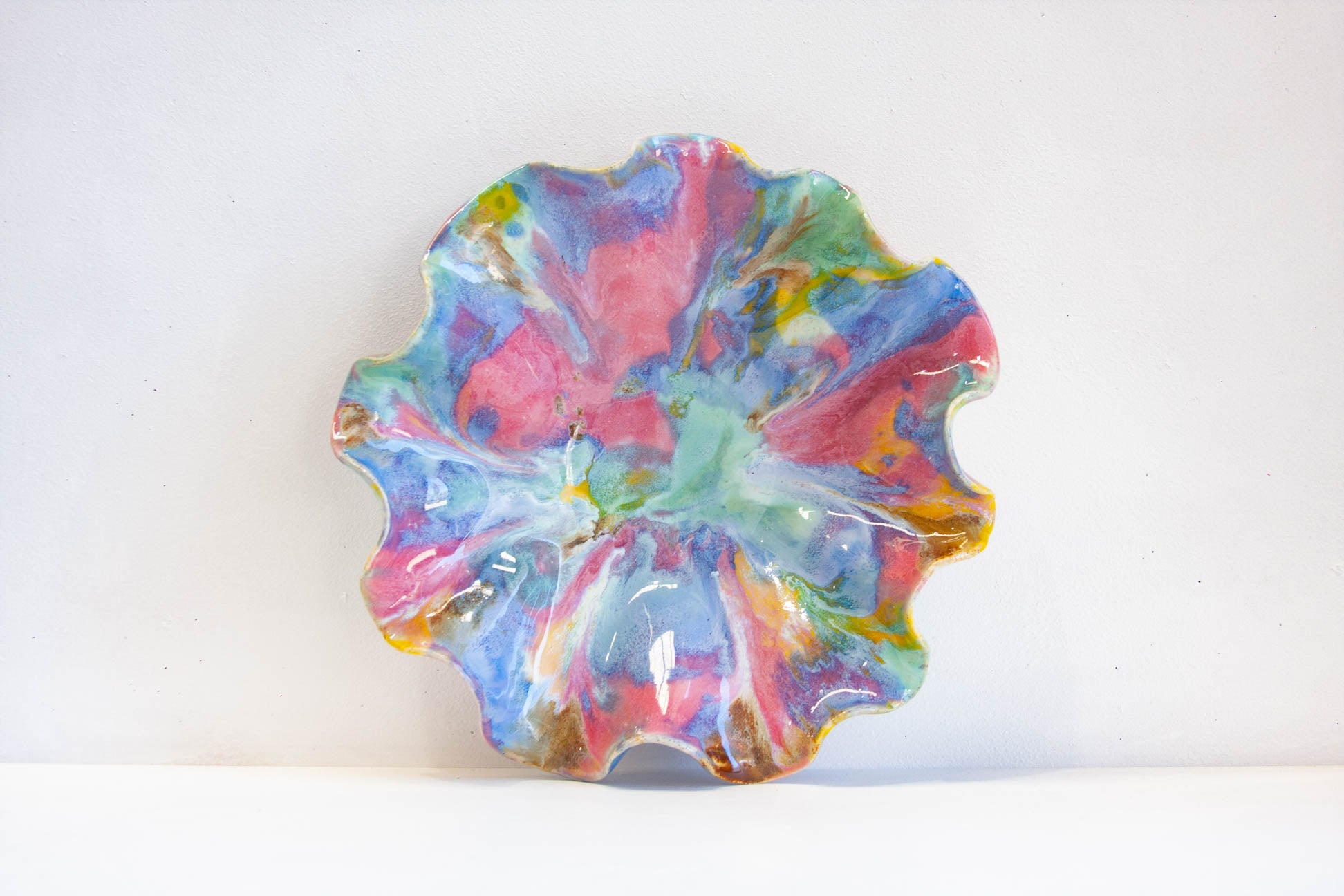 A handmade ceramic plate with a wavy, scalloped rim, decorated with a vibrant, multicolored marbled glaze of pink, blue, green, yellow, and white on a plain white background.