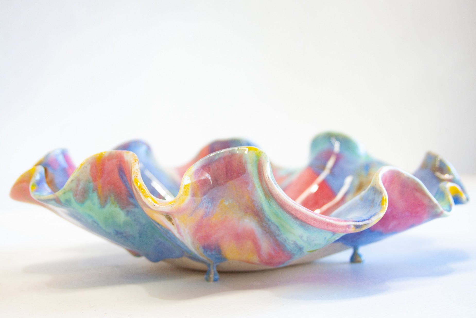 A colorful, handcrafted ceramic bowl with a wavy, ruffled rim and small feet, decorated with a marbled pattern of pink, blue, yellow, and green glaze, set against a plain white background.