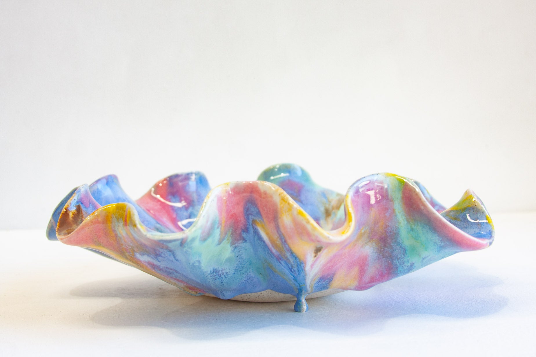 A handmade ceramic bowl with a wavy, ruffled rim, decorated in a marbled swirl pattern of pastel pink, blue, yellow, and green glaze, set against a plain white background.