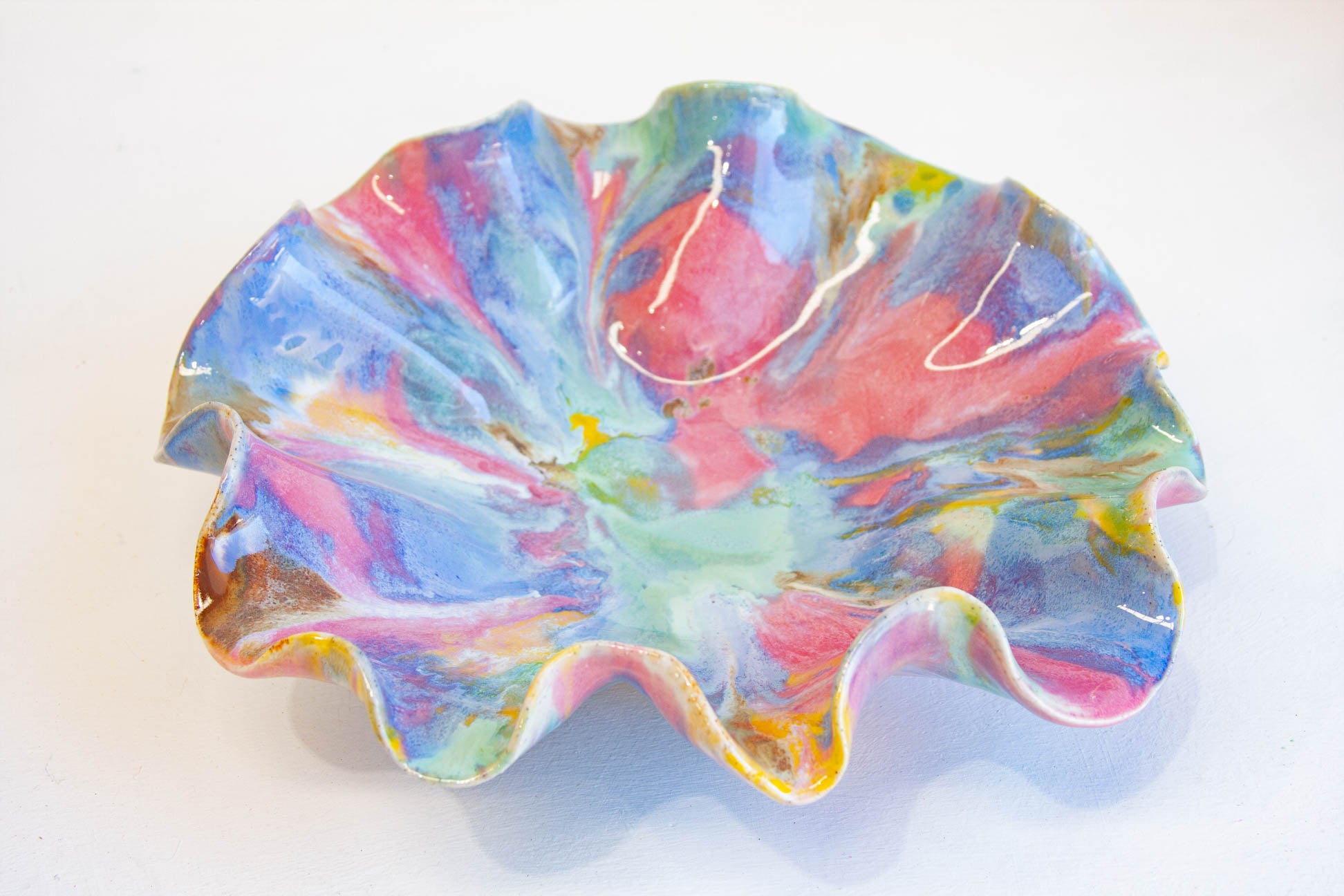 A handmade ceramic dish with a wavy, ruffled edge, decorated with a vibrant, marbled swirl pattern of pink, blue, green, yellow, and white glaze on a white background.