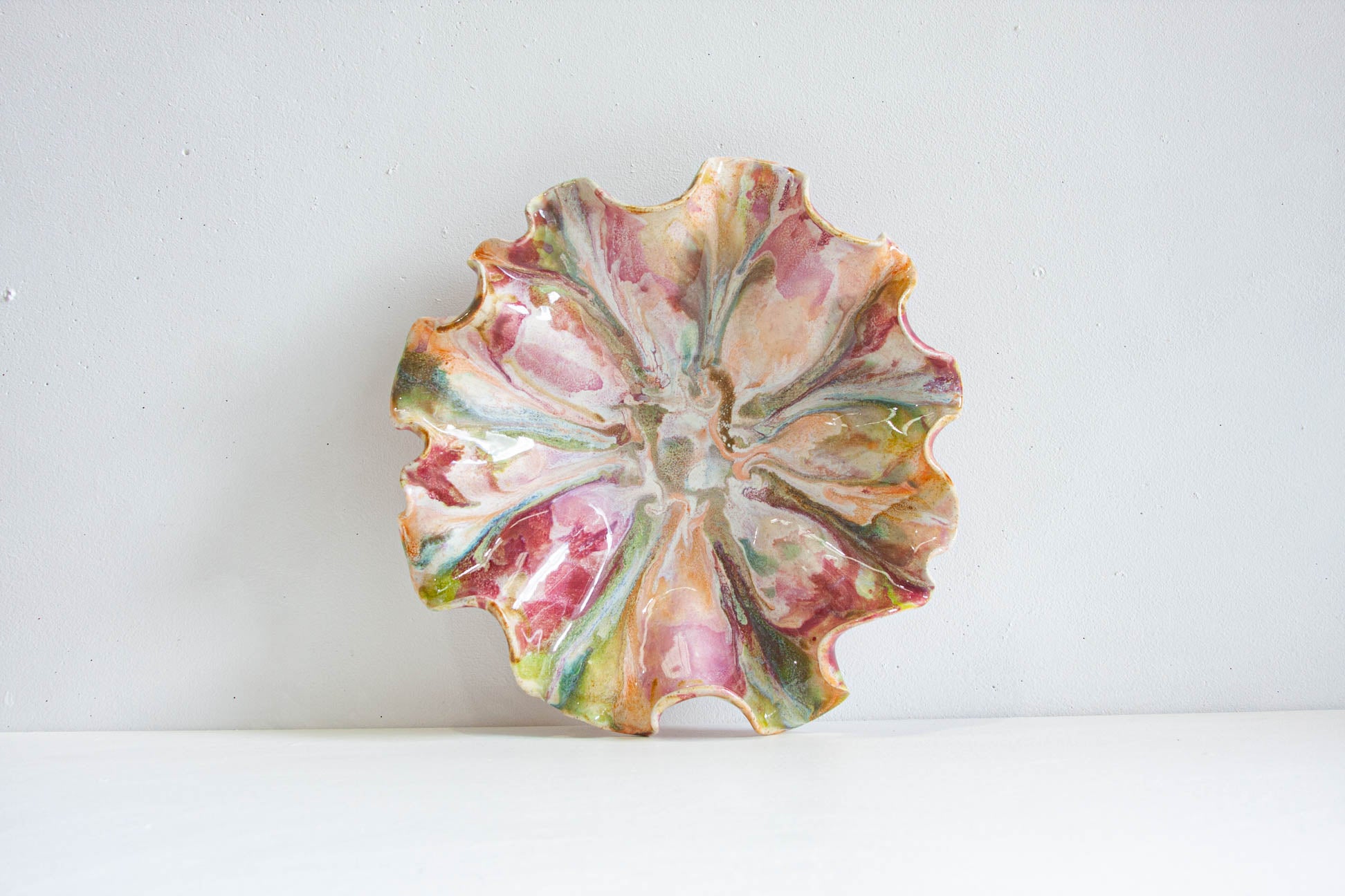 A scalloped ceramic plate with a marbled pattern of pink, green, orange, and cream glazes, displayed against a plain white wall and surface.