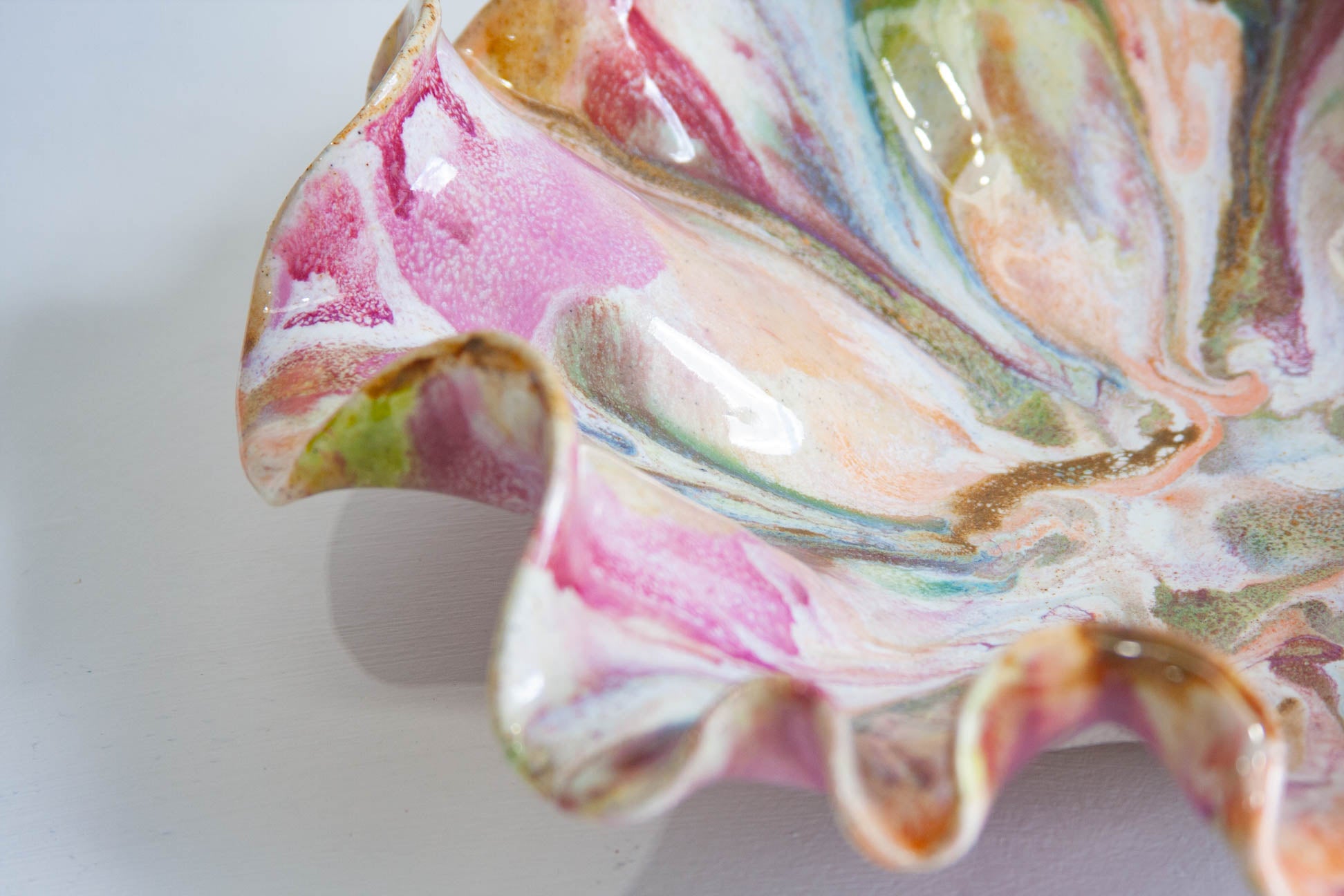 A close-up of a decorative ceramic dish with a ruffled edge, featuring a marbled pattern of pink, green, blue, and gold glazes on a white base.