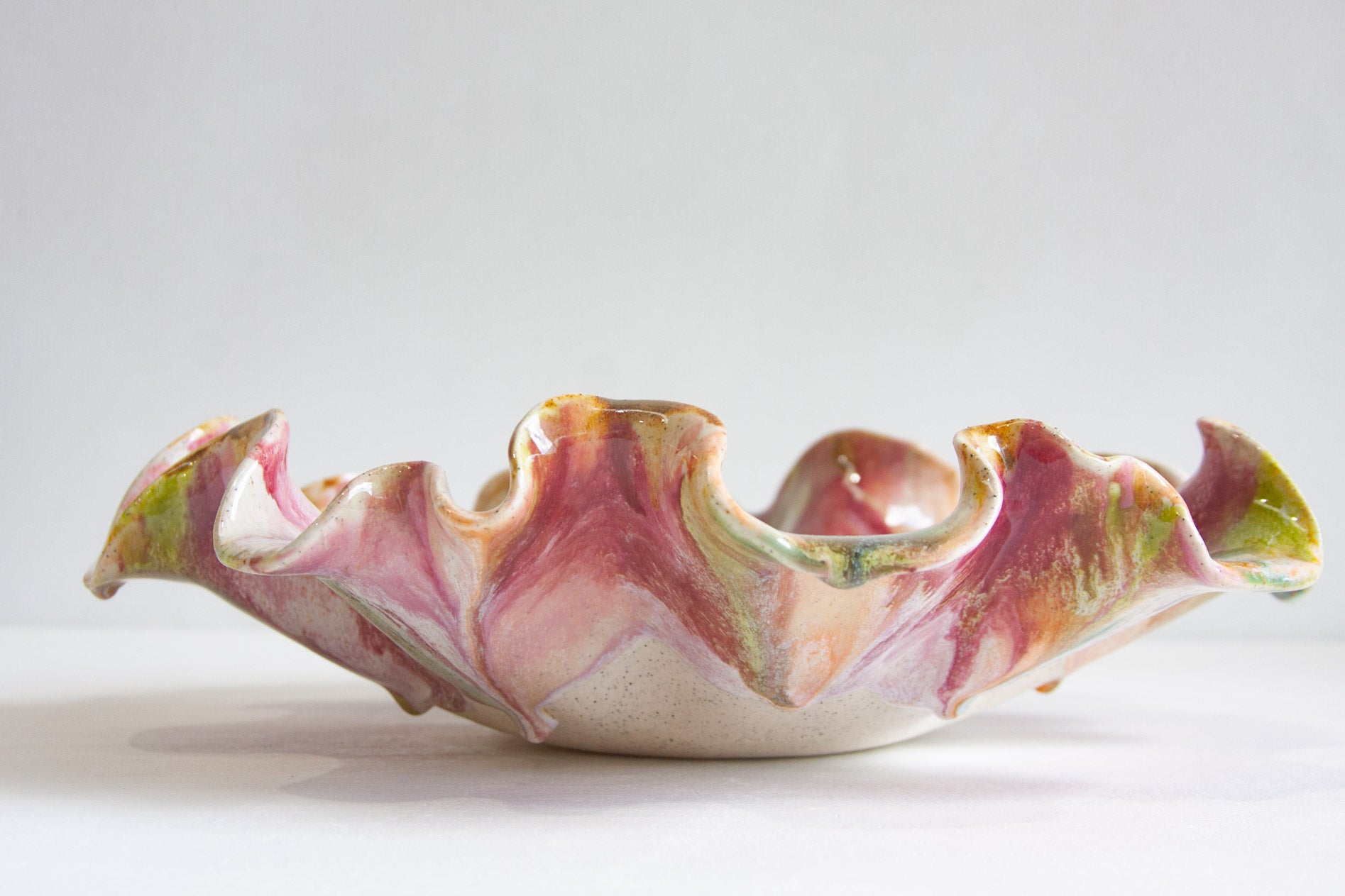 A handmade ceramic bowl with a ruffled, petal-like edge, featuring a marbled glaze of pink, green, and gold over a white base, photographed on a light gray background.