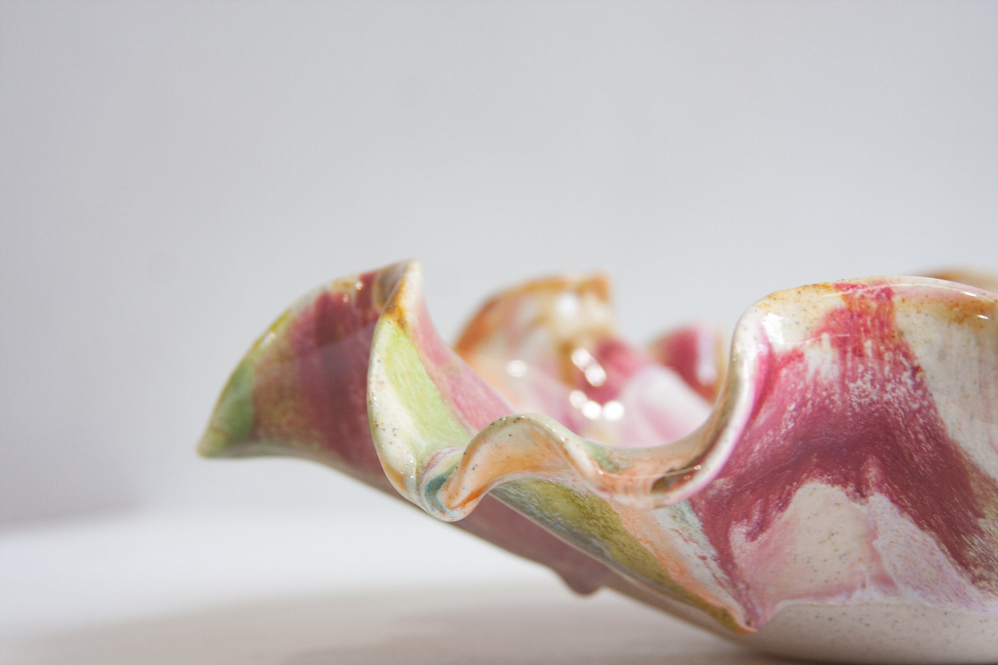 A close-up of a wavy ceramic dish with a glossy finish, featuring marbled pink, green, and cream glazes on a light gray background.