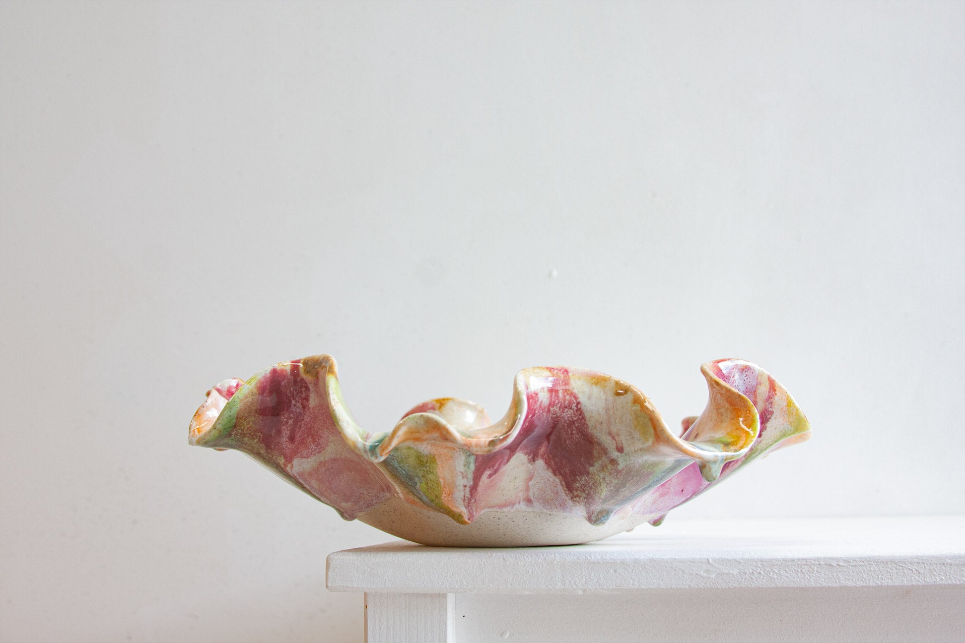 A handmade ceramic bowl with a wavy, ruffled rim, decorated in a marbled glaze of pink, green, yellow, and white, sitting on a white table against a white wall.
