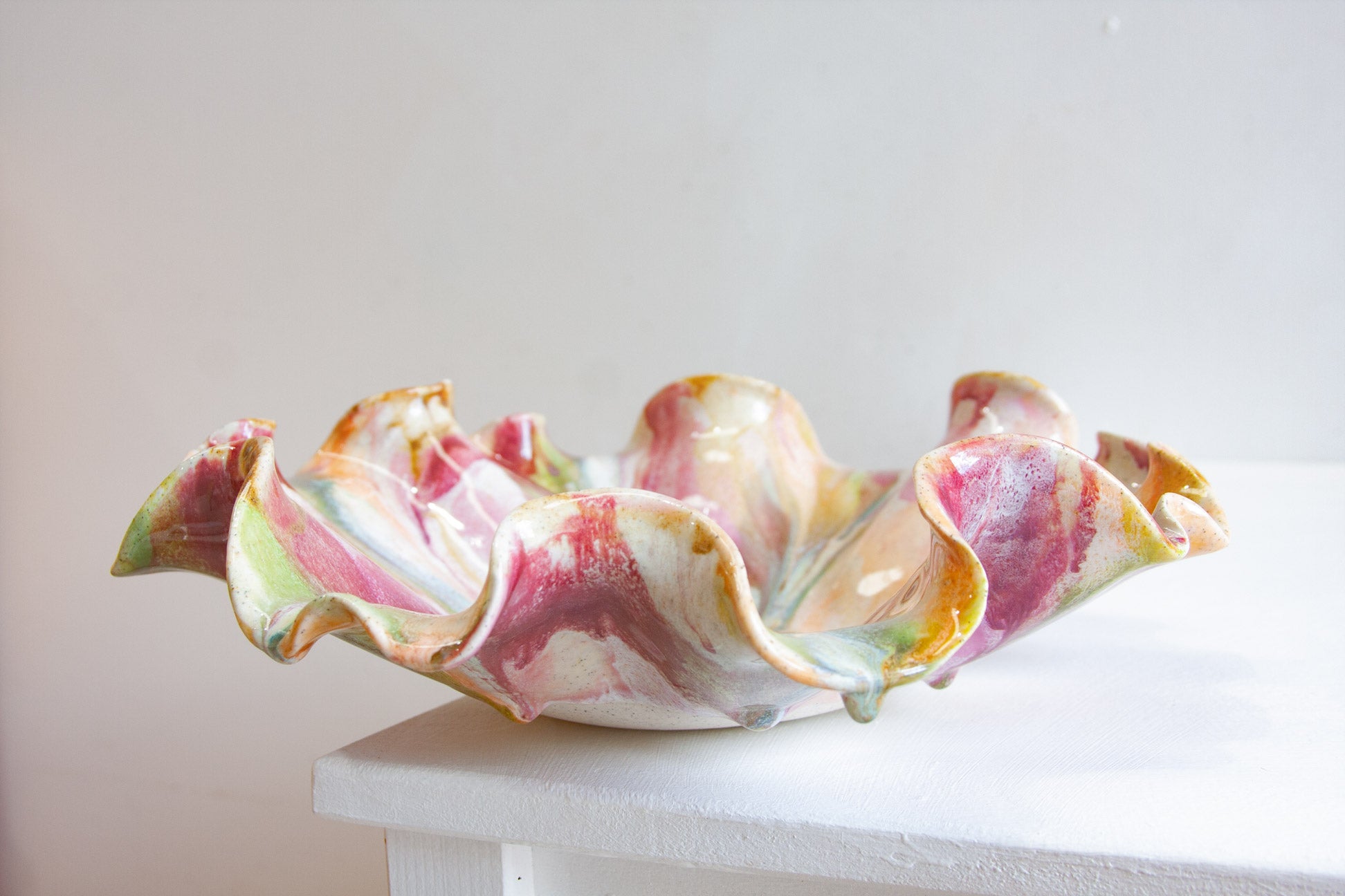 A handmade ceramic flower-shaped bowl with ruffled petals, featuring a marbled glaze of pink, green, yellow, and white, displayed on a white table against a neutral light gray background.