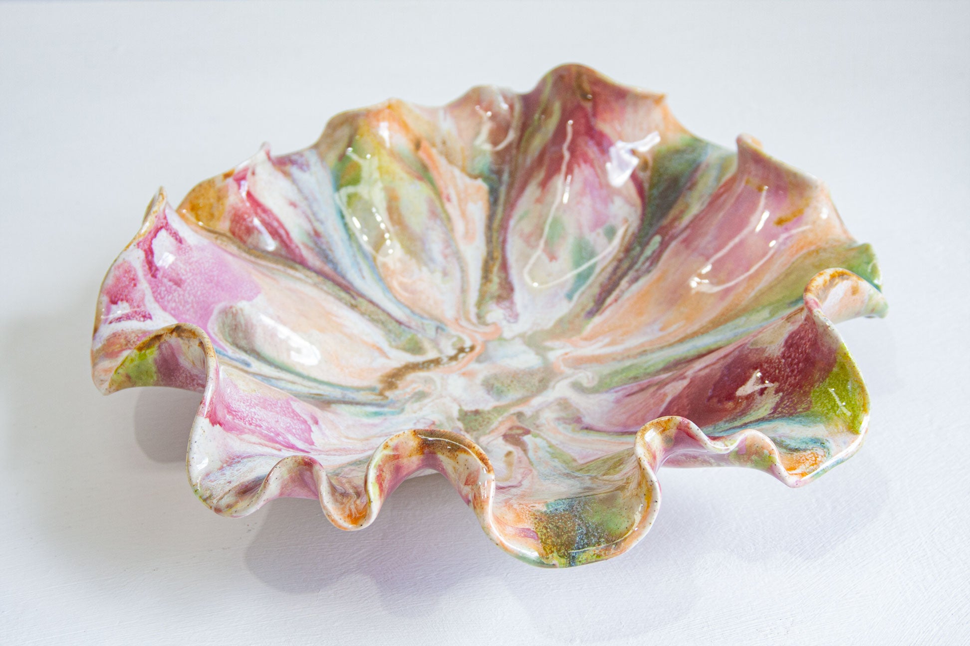 A handmade ceramic dish with a ruffled, lotus-like edge, decorated with a marbled glaze of pink, green, orange, and white on a white background.
