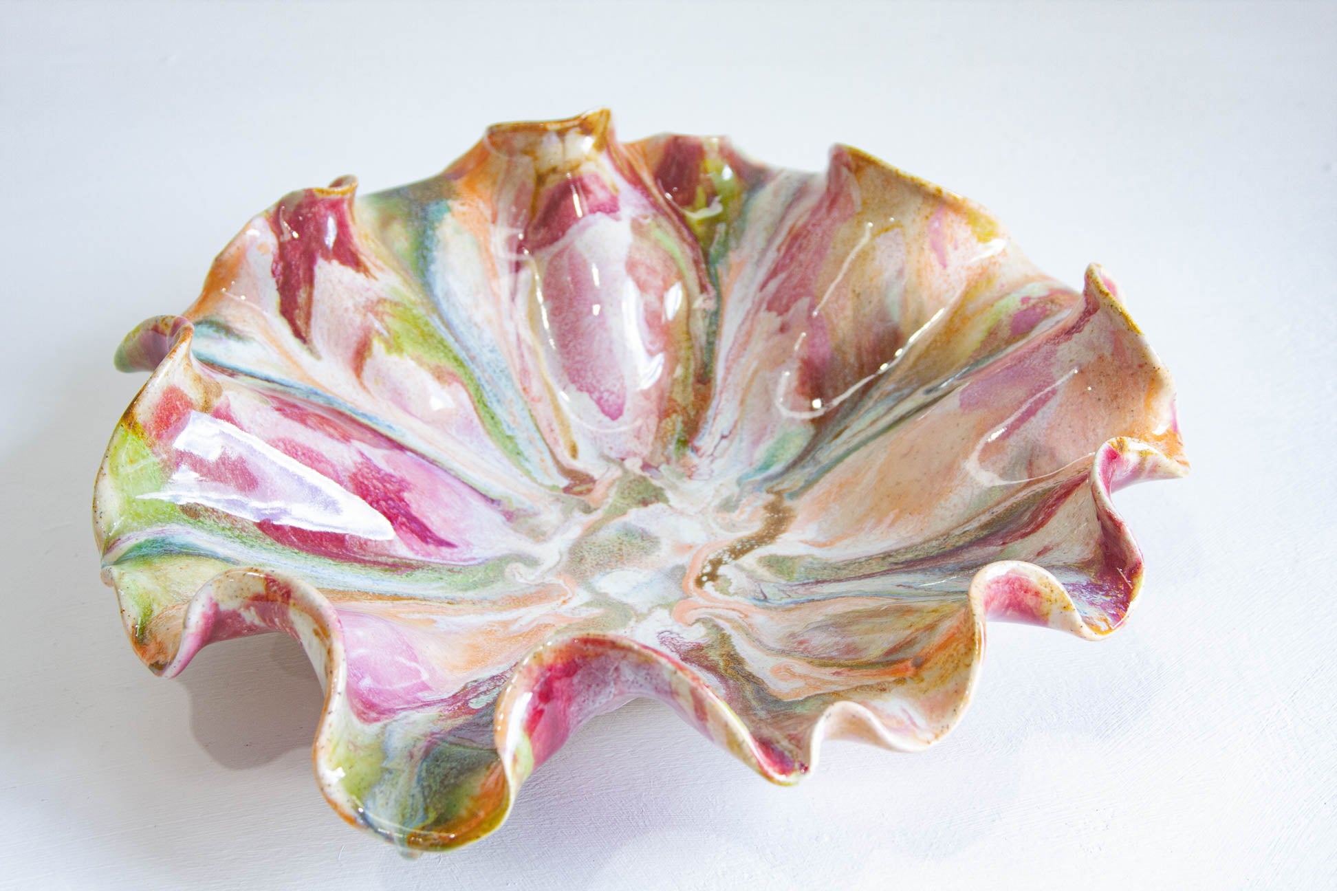 A handmade ceramic dish with a ruffled, petal-like edge, decorated with a marbled glaze of pink, green, gold, and white, shown on a plain white background.