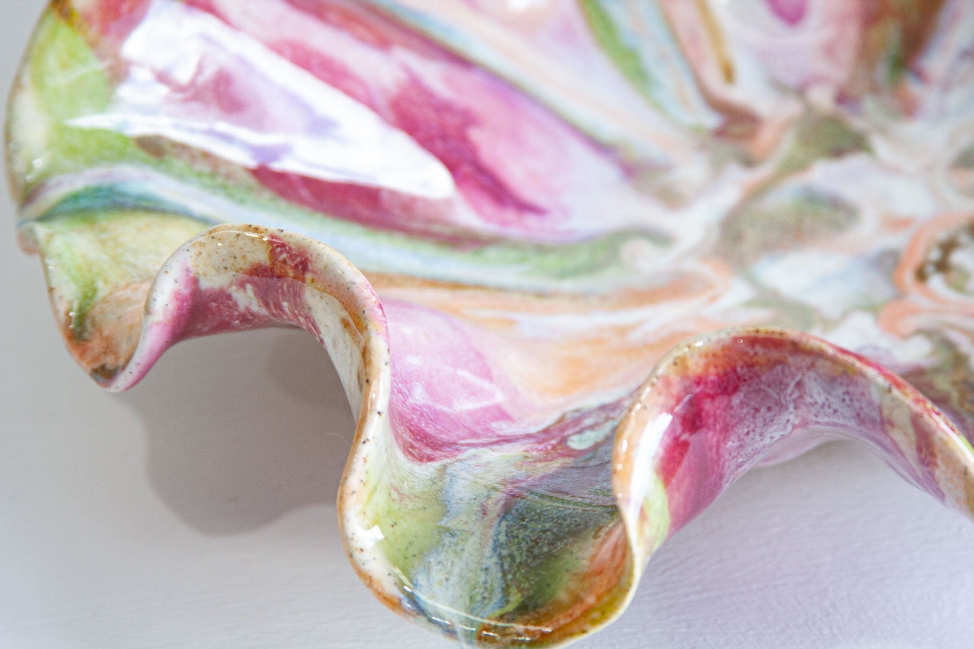 Close-up of a ceramic plate with a wavy rim, decorated with a marbled pattern of pink, green, orange, and white glazes on a white background.