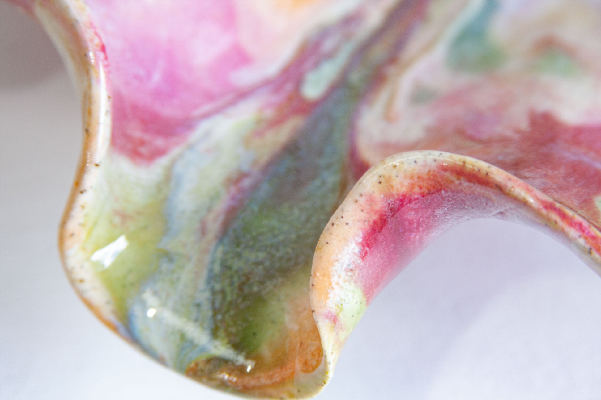 Close-up of a glossy ceramic bowl with a wavy rim, featuring a marbled glaze in pink, green, blue, and beige tones.