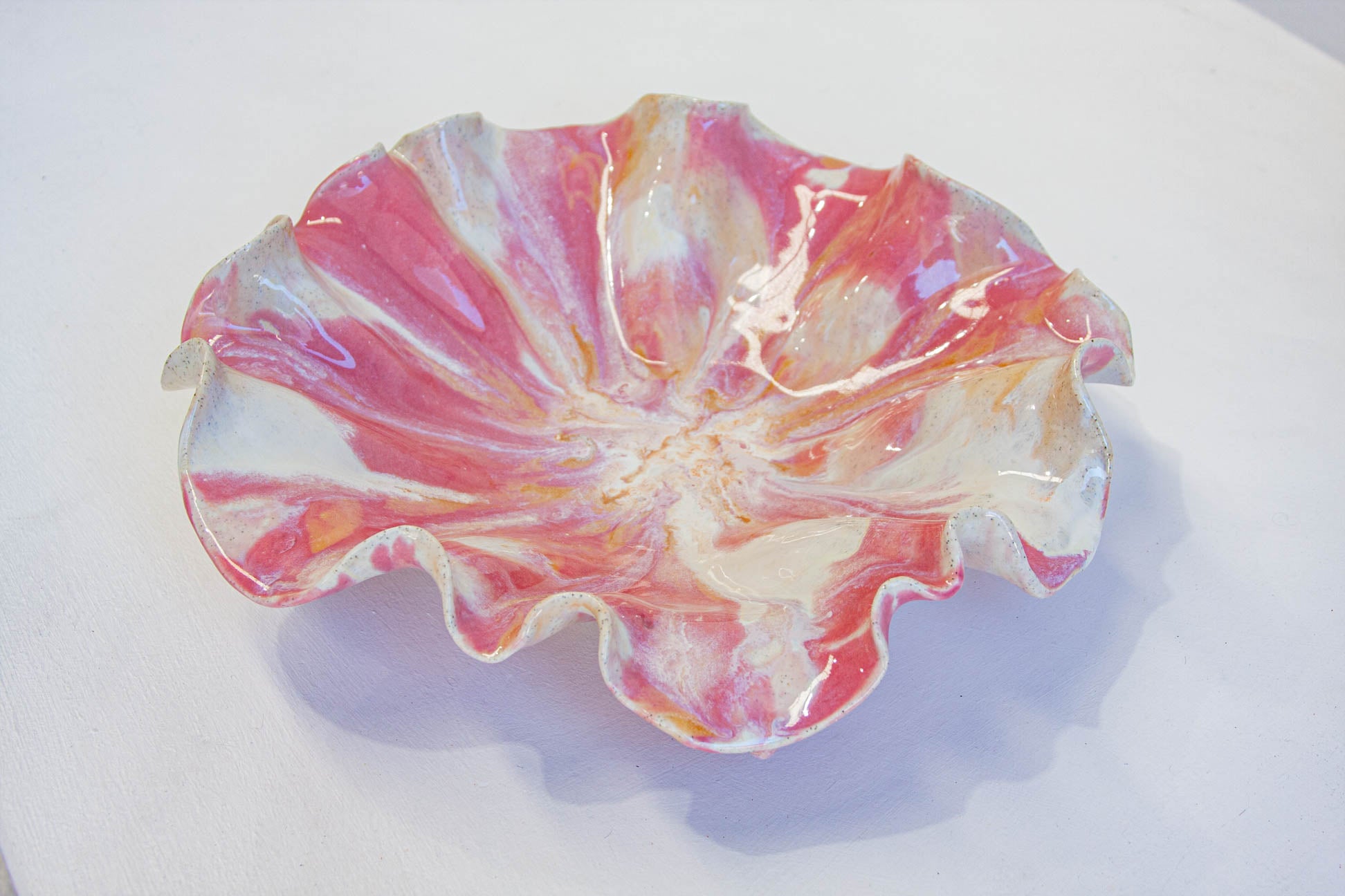 A handmade ceramic bowl with a ruffled, petal-like edge, decorated with a marbled pattern of pink, red, orange, and white glaze, shown on a plain white surface.