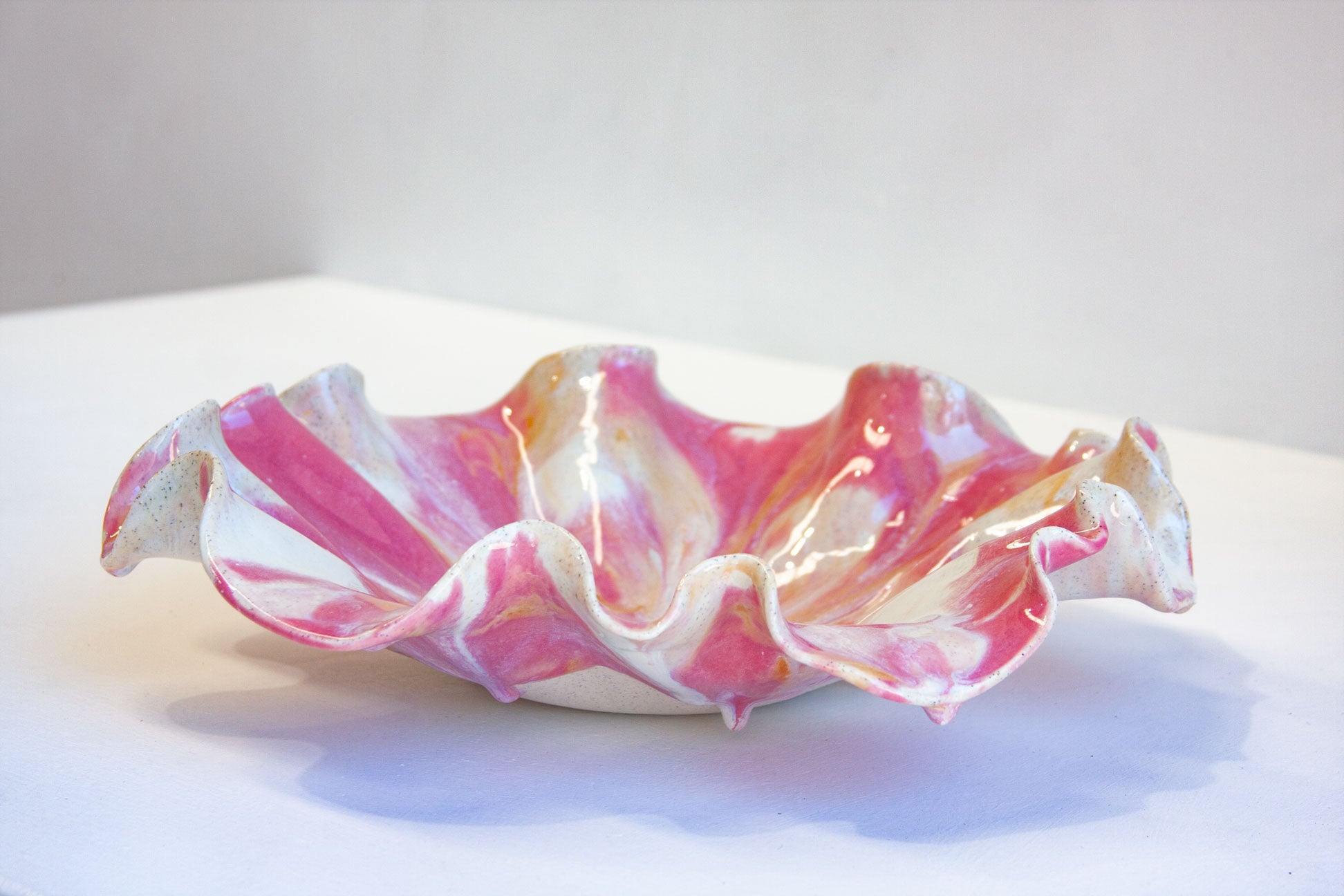 A handmade ceramic ruffled bowl with a marbled pattern of pink, white, and subtle gold glaze, resting on a white surface against a light gray background.