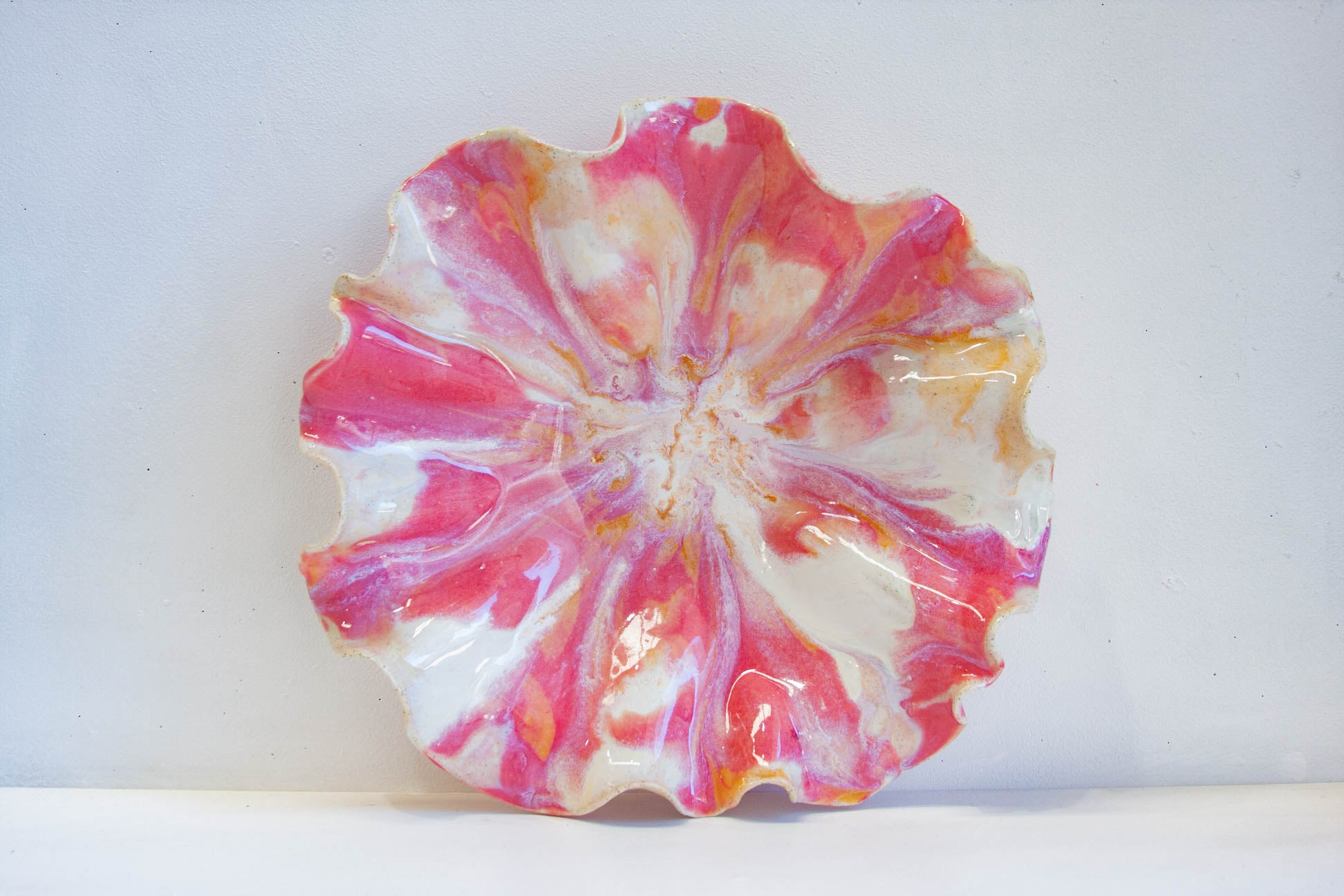 A round, scalloped-edge ceramic plate with a vibrant pink, white, and orange marbled pattern, photographed against a plain white background.