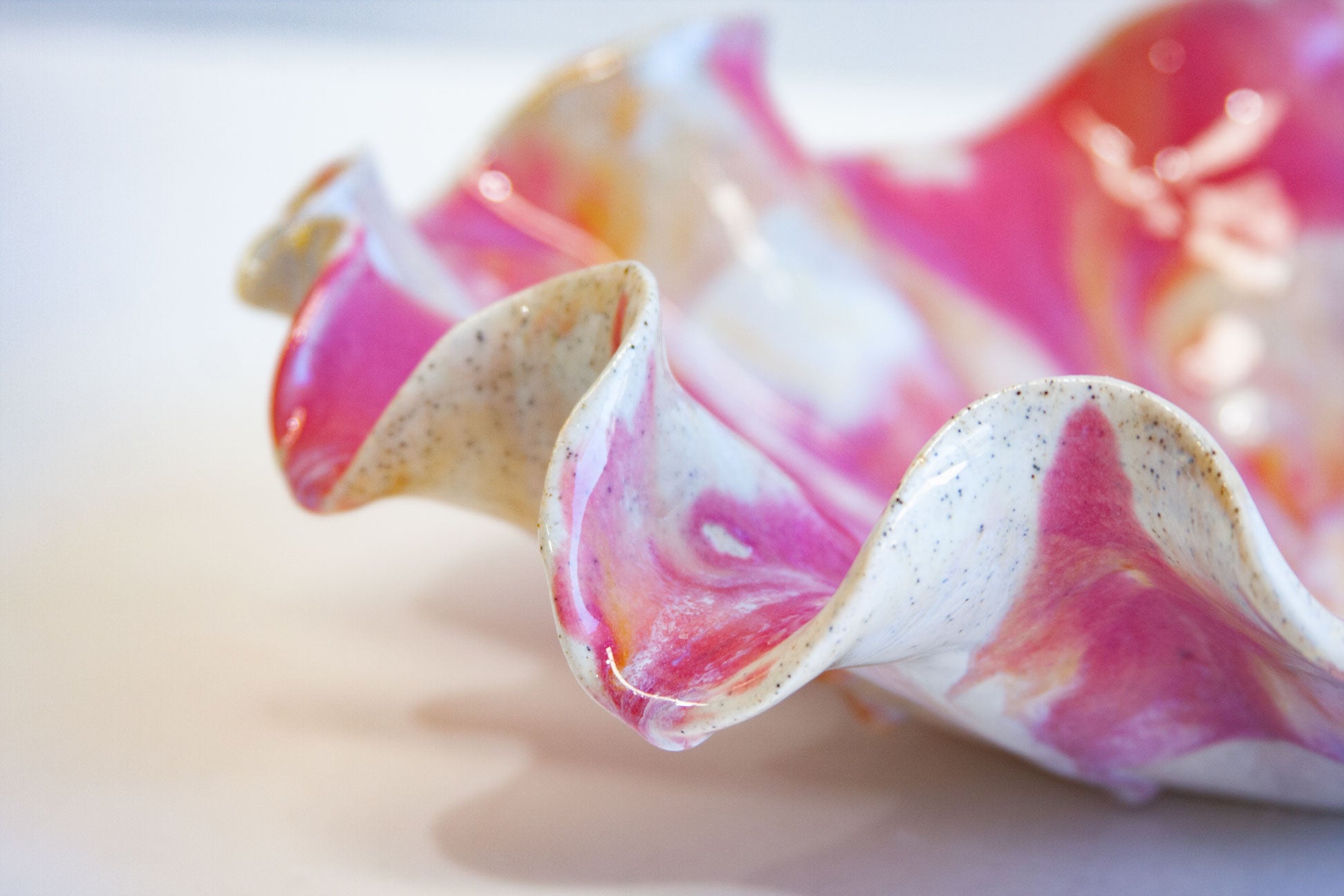 A close-up of a glossy ceramic shell-shaped vessel with wavy, fluted edges, featuring a marbled pattern of vibrant pink, white, and subtle gold speckles on a light background.