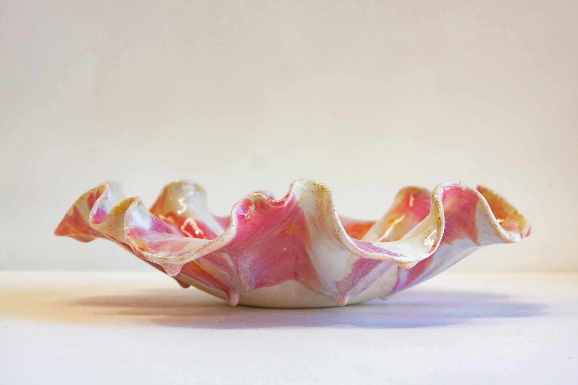 A handmade ceramic bowl with a wavy, ruffled rim, featuring a marbled pattern of pink, white, and subtle gold glaze, set against a plain light beige background.