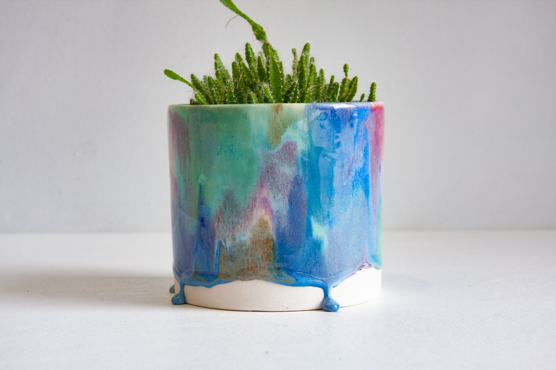 A small potted succulent in a handmade hexagonal ceramic planter with a colorful blue, green, and purple drip glaze, set against a plain white background.