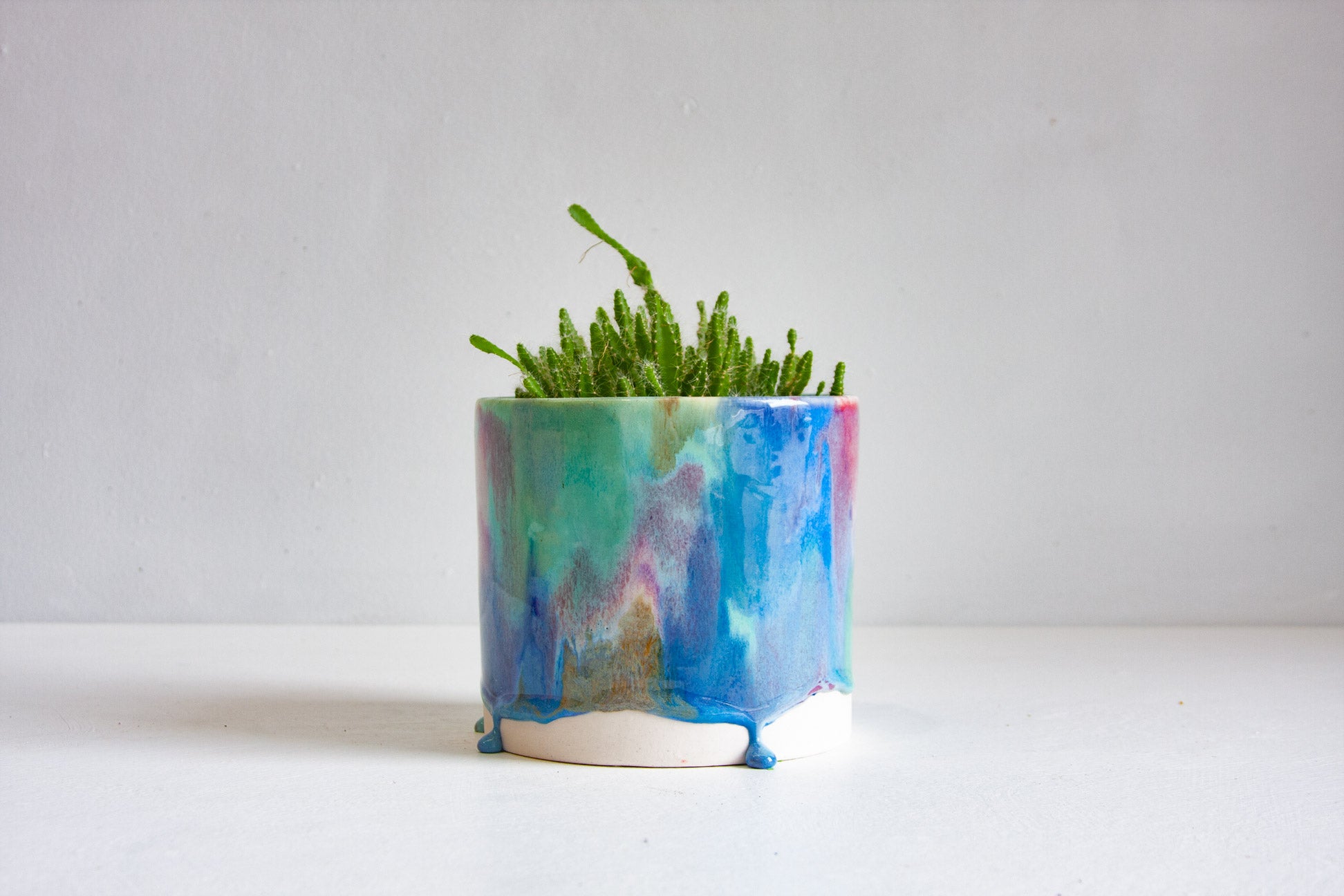 A small green succulent plant in a handmade ceramic pot with a marbled blue, teal, and purple glaze, set against a plain light gray background.