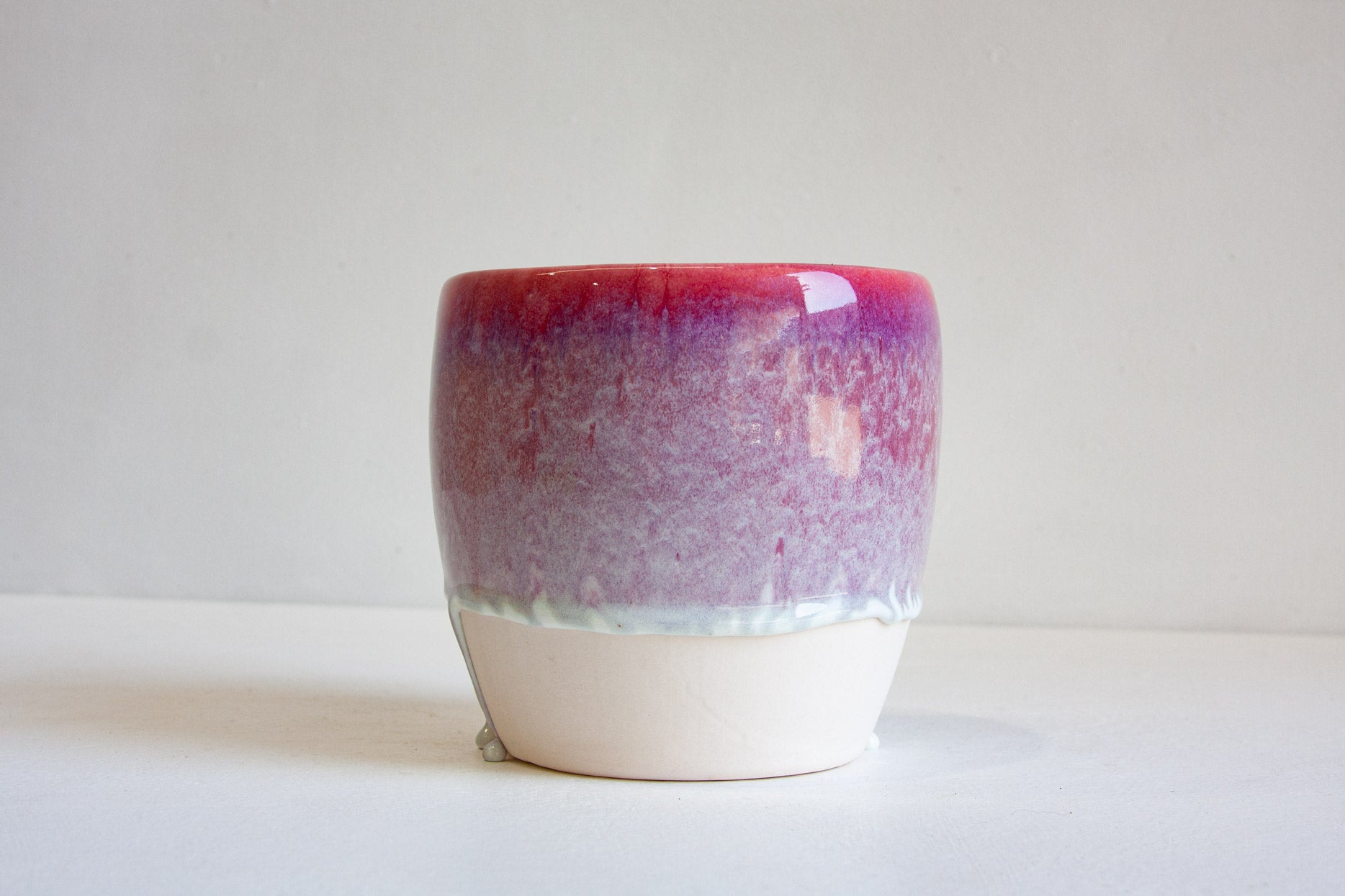 A handmade ceramic cup with a glossy gradient glaze transitioning from deep magenta to lavender-purple, finished with a matte off-white base, set against a plain light gray background.