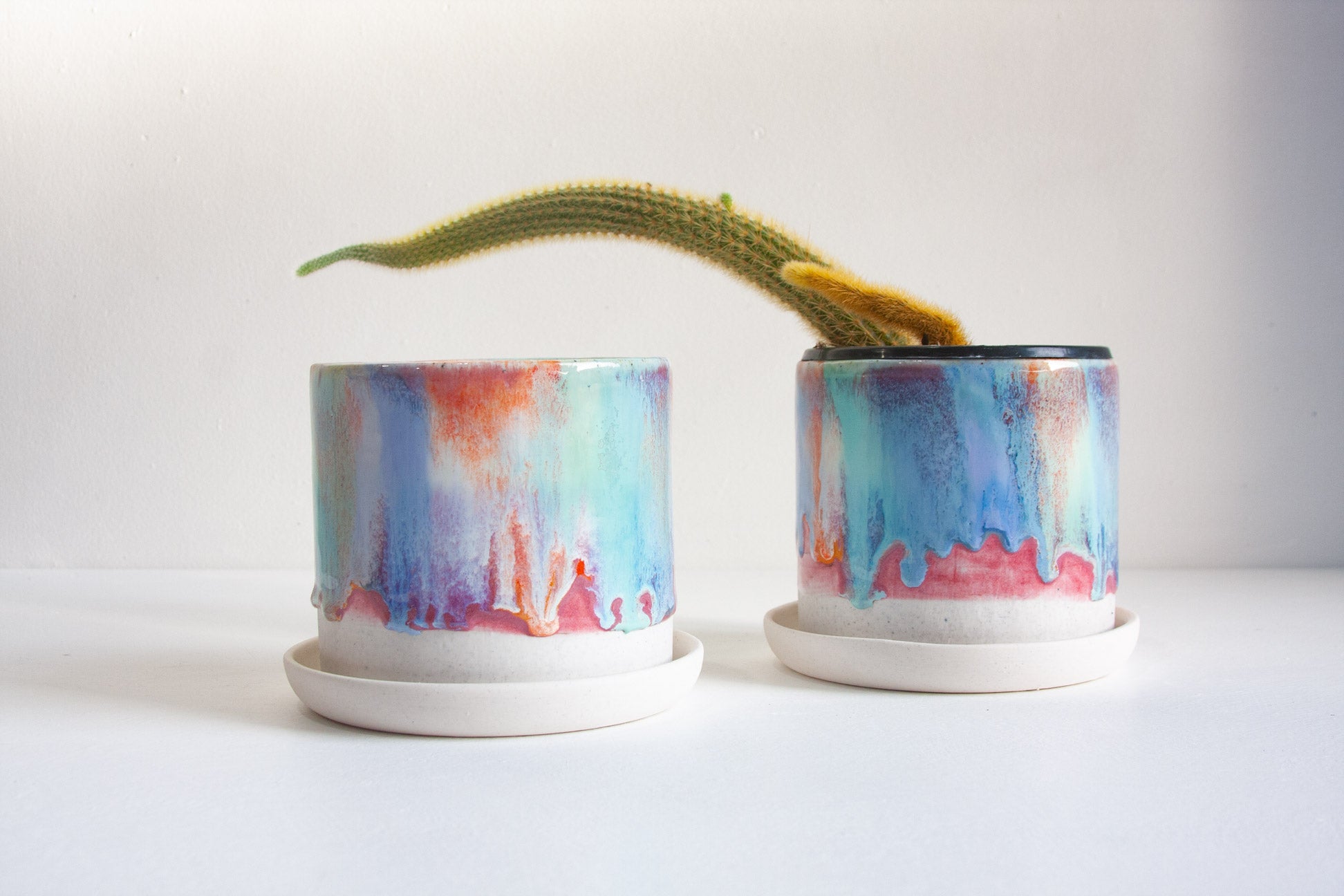Two handmade ceramic multicoloured planters, one contains a cactus and one is empty, both sitting on ceramic saucers.