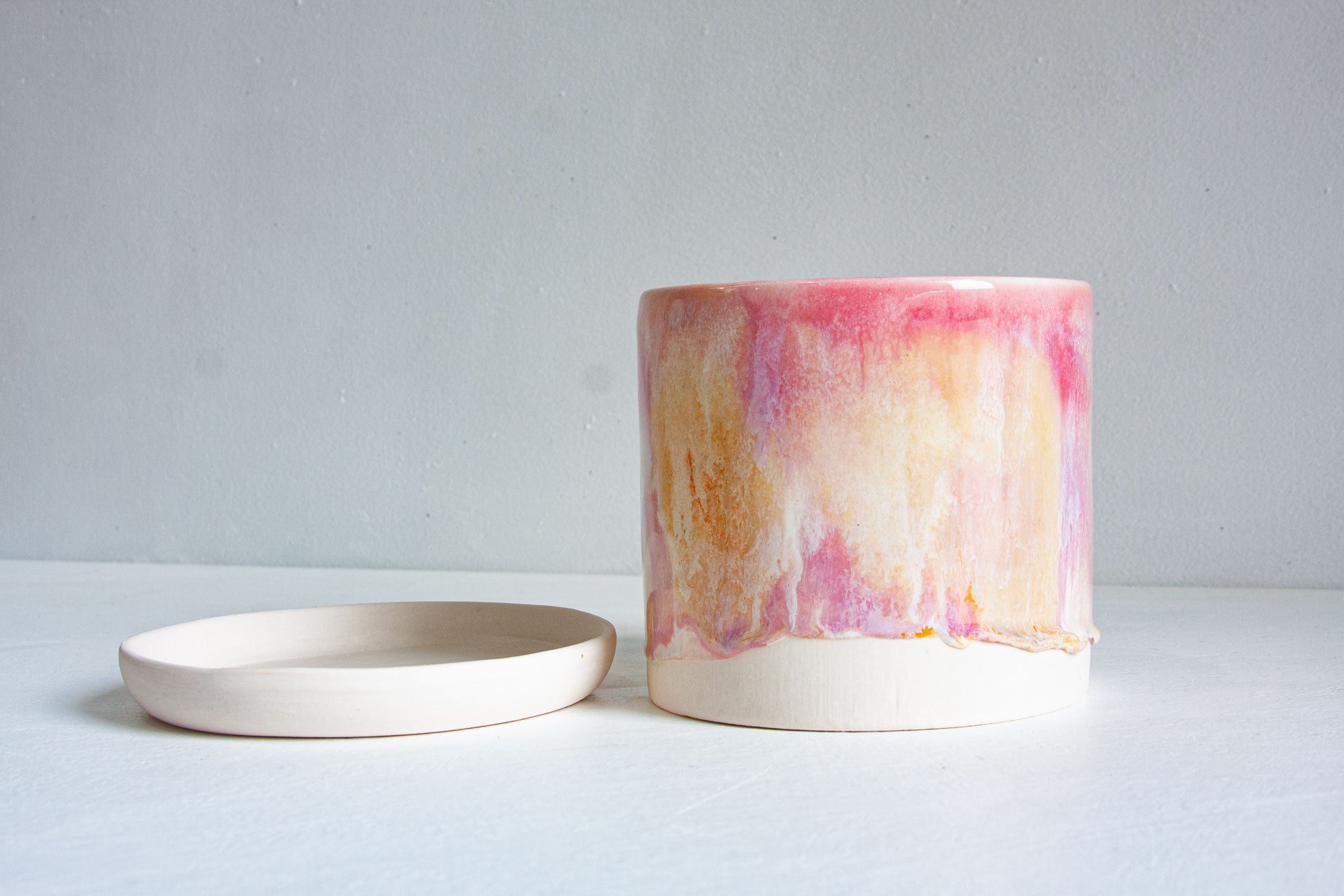 A cylindrical ceramic planter with a dripping pink, peach, and cream glazed finish, shown next to its matching shallow saucer on a light gray surface.