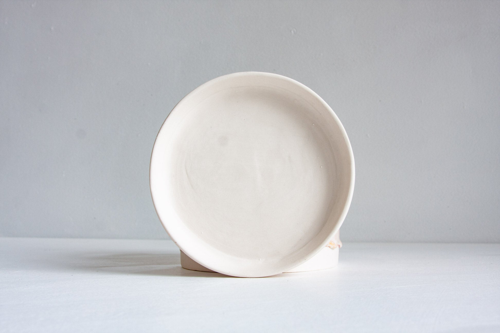 A minimalist ceramic plate in a warm off-white or cream glaze, displayed on a small wooden riser against a clean, light gray studio background.