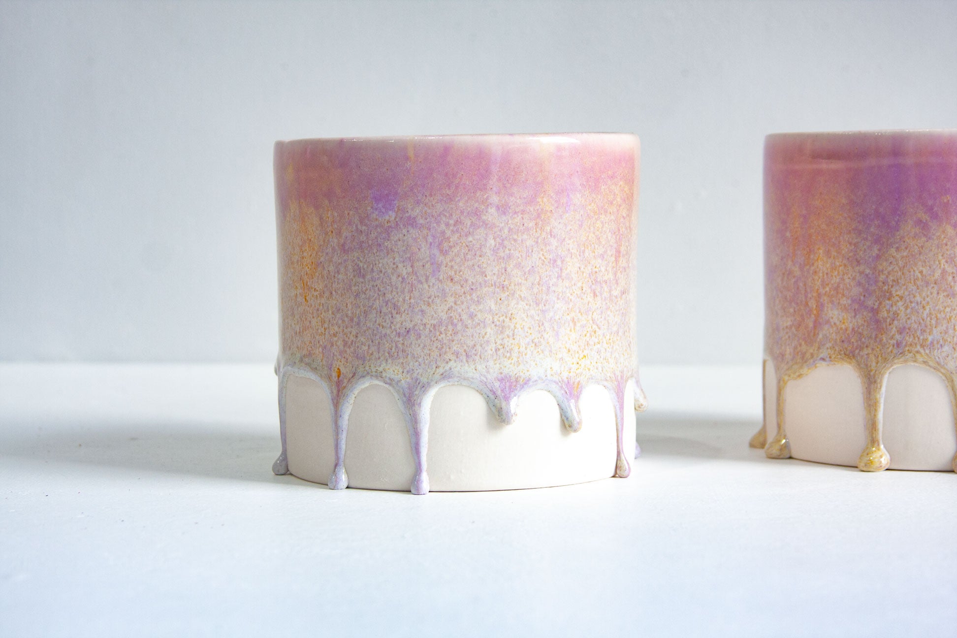 Two ceramic drip pots with a pink, purple, and gold iridescent glaze on the upper half and a matte off-white base, shown on a white surface against a light grey background.
