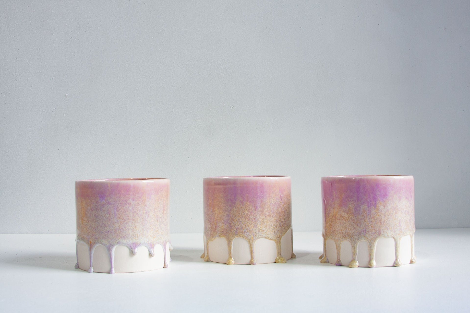 Three small cylindrical ceramic vases with a pink, purple, and gold marbled glaze and white drip detailing at the base, arranged on a white surface against a light grey background.