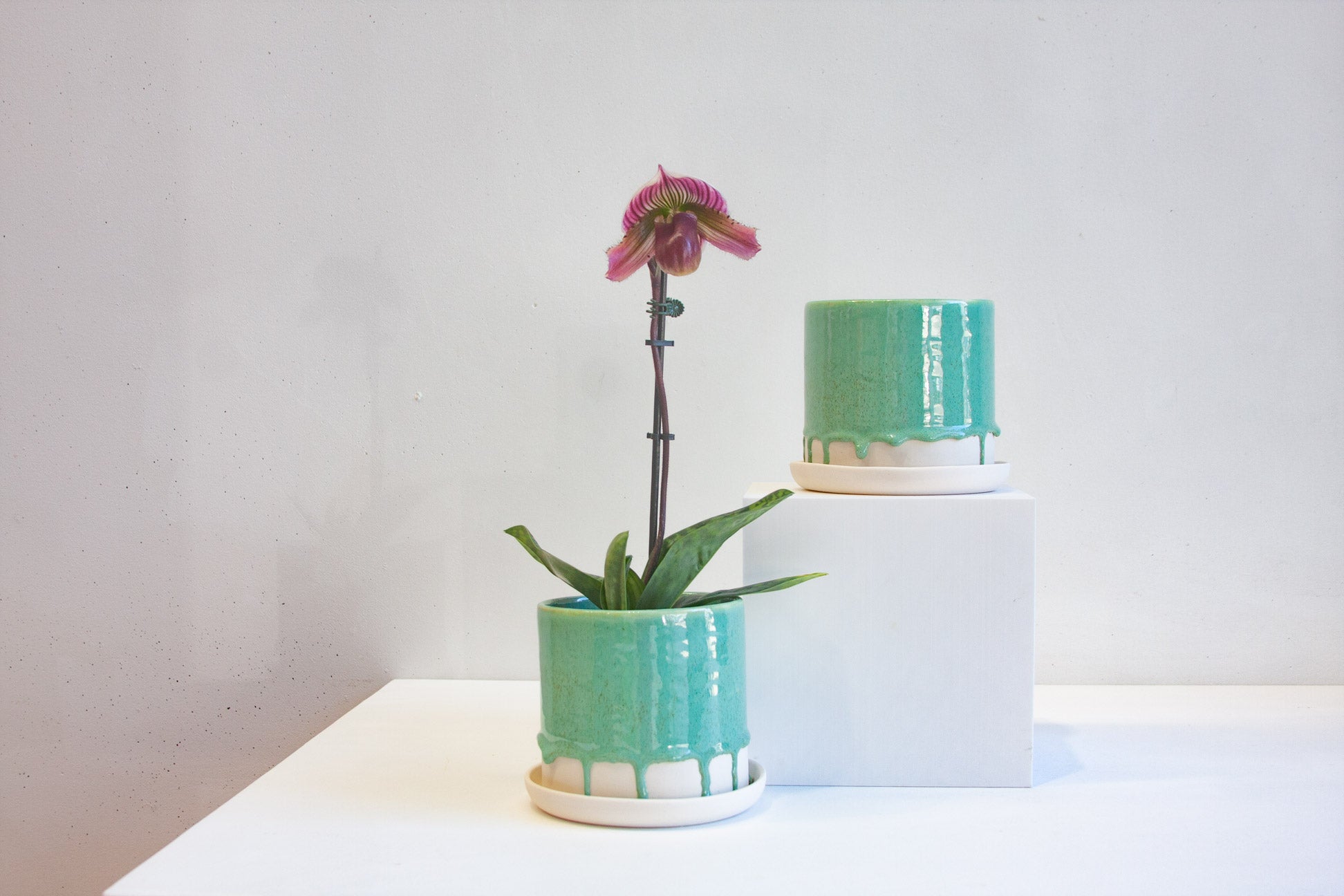 two handmade ceramic drippy green planter, one sits on a white plinth the other beside it on the table.