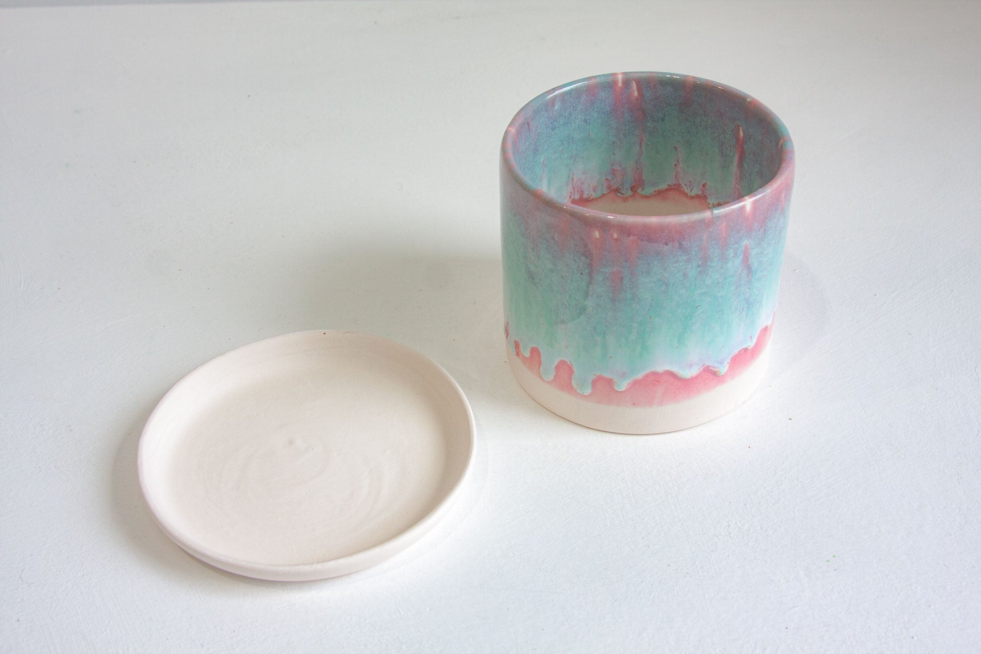 A ceramic cup with a matching small round saucer, featuring a dripping glaze design in teal, pink, and cream on a white background.