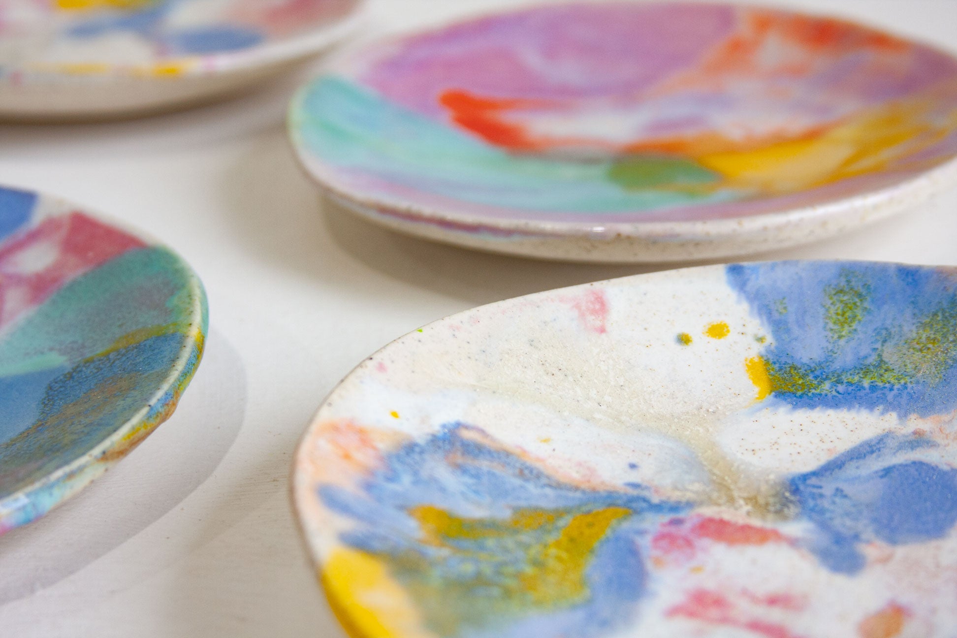 Four handmade ceramic small plates arranged on a white surface, glazed in pink, blue, yellow, orange and green over speckled clay.