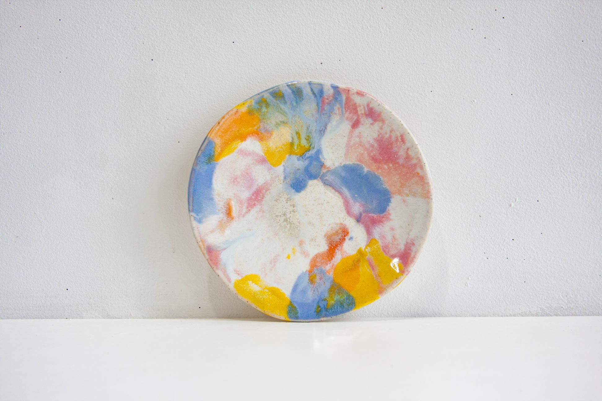 A handmade ceramic small plate in the Daisy colourway, yellow, blue, pink and peach washing across speckled white, propped against a pale wall.