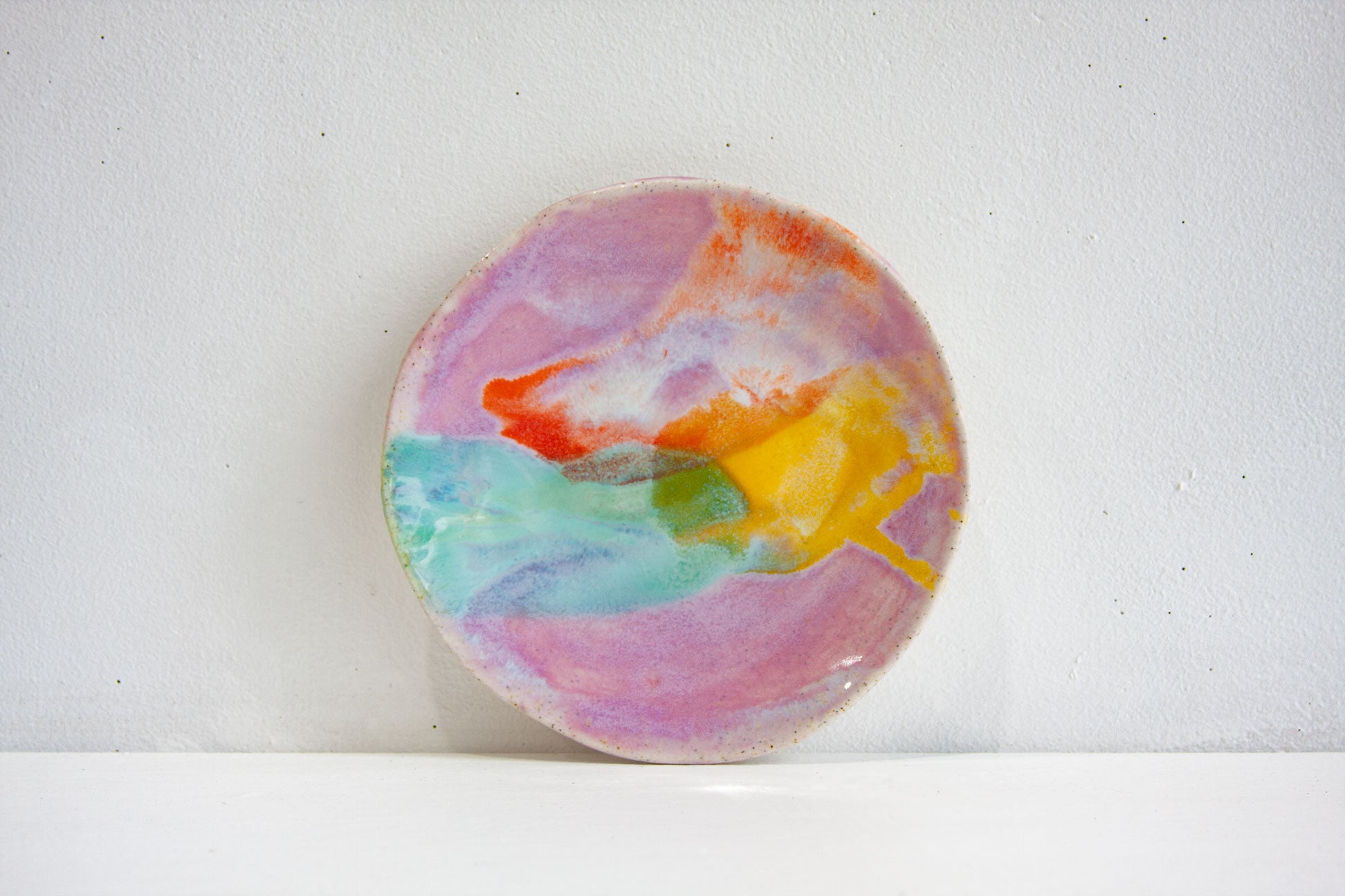 A handmade ceramic small plate in the Colour Theory colourway, lilac, orange, yellow and green running into one another, propped against a pale wall.