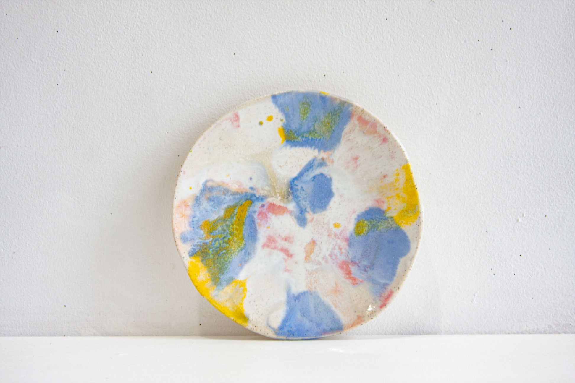 A handmade ceramic small plate in the Daisy colourway, blue and yellow with pink and peach over speckled white, propped against a pale wall.