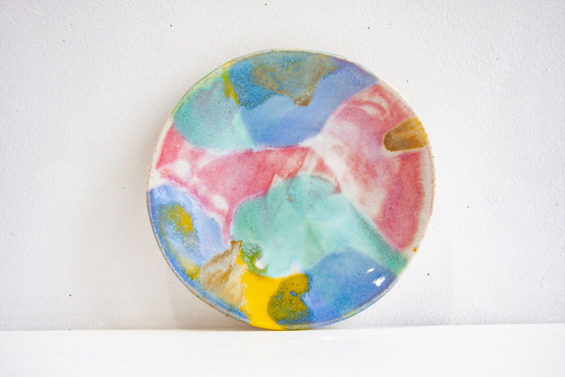 A handmade ceramic small plate in the Rainbow colourway, pink, blue, green and yellow meeting across the surface, propped against a pale wall.