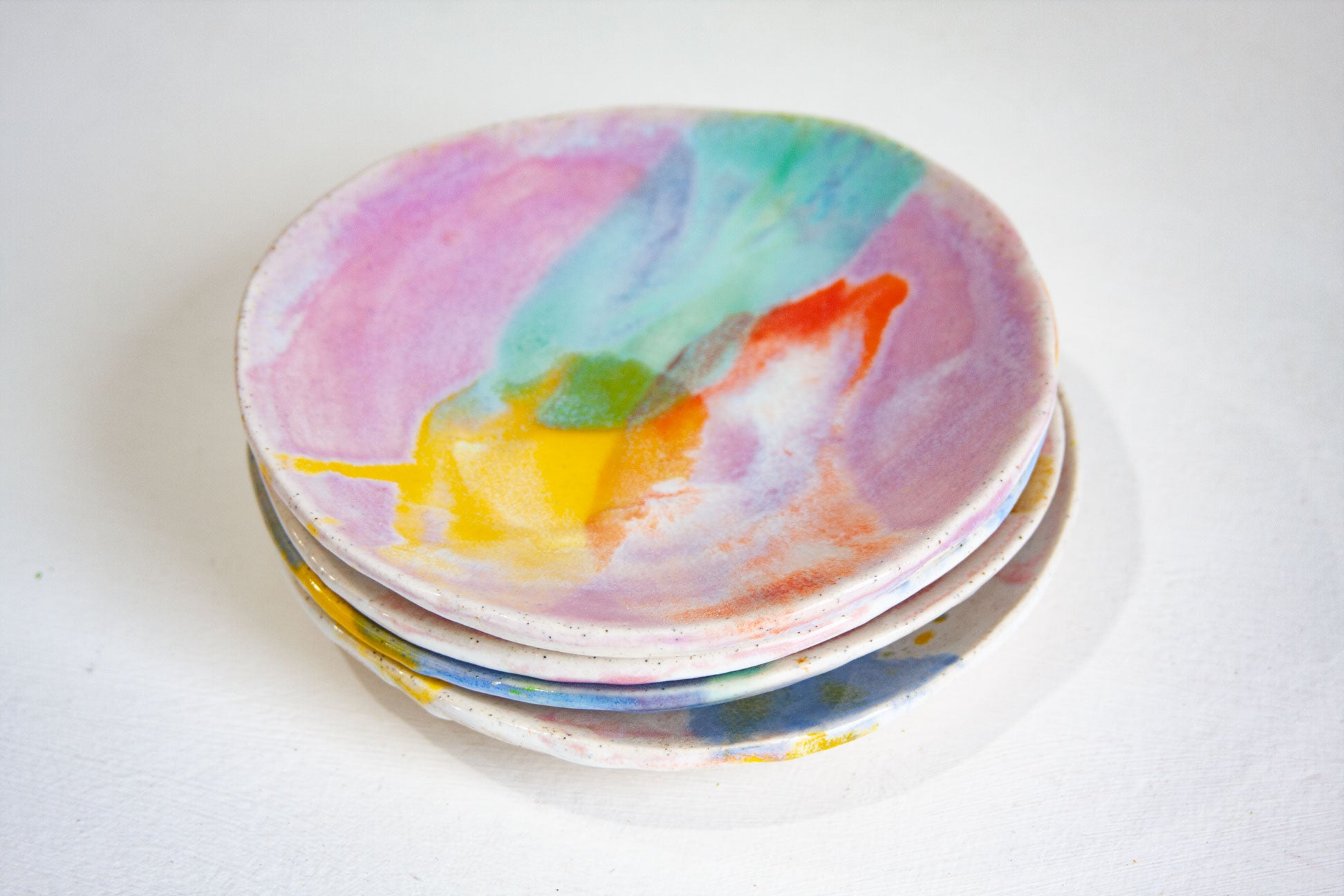 Four handmade ceramic small plates stacked, the top one in Colour Theory with lilac, green, orange and yellow, the others showing at the edges.