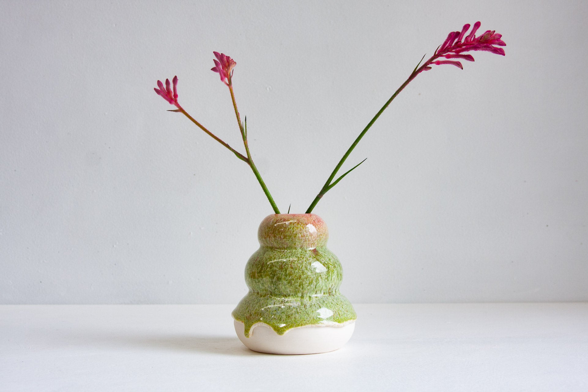 A small handmade ceramic vase with a textured green and white body and a pinkish-orange glazed rim, holding two long green stems with magenta flowers, set against a plain light gray background.