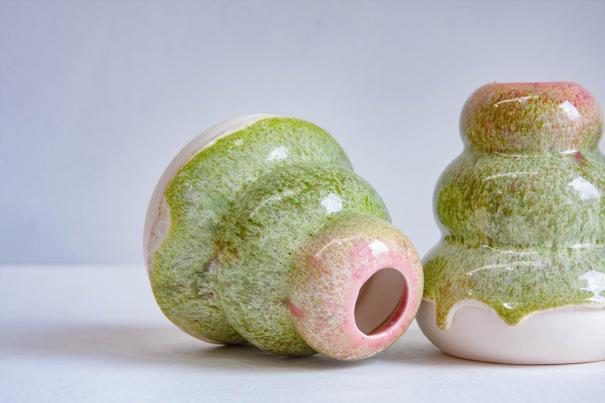 Two small glossy ceramic vessels with a green and pink marbled glaze, one standing upright and one lying on its side, photographed on a light gray studio background.