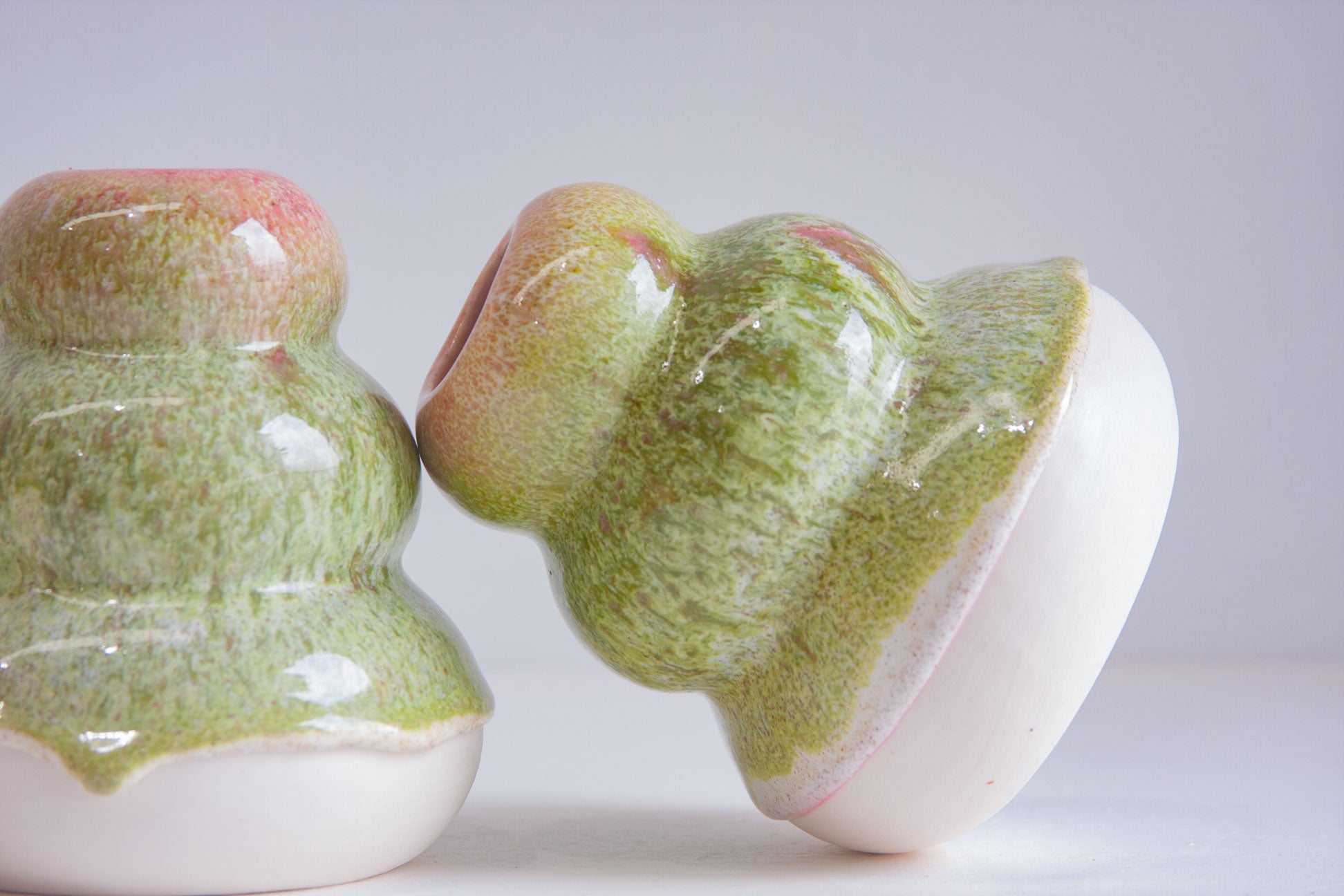 Two small glossy ceramic objects with a green mottled glaze and white bases, photographed on a light gray studio background.