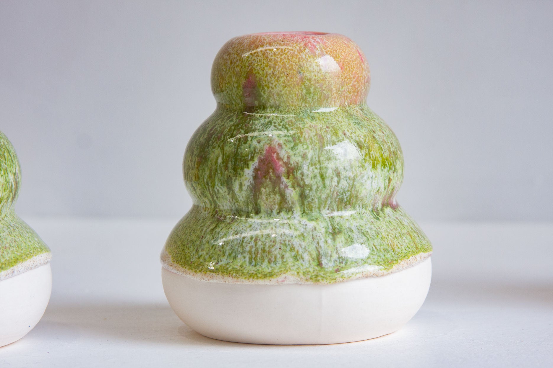 A small ceramic bud vase with a glossy green and pink glaze, featuring a rounded tiered body and a white base, shown on a light gray background.